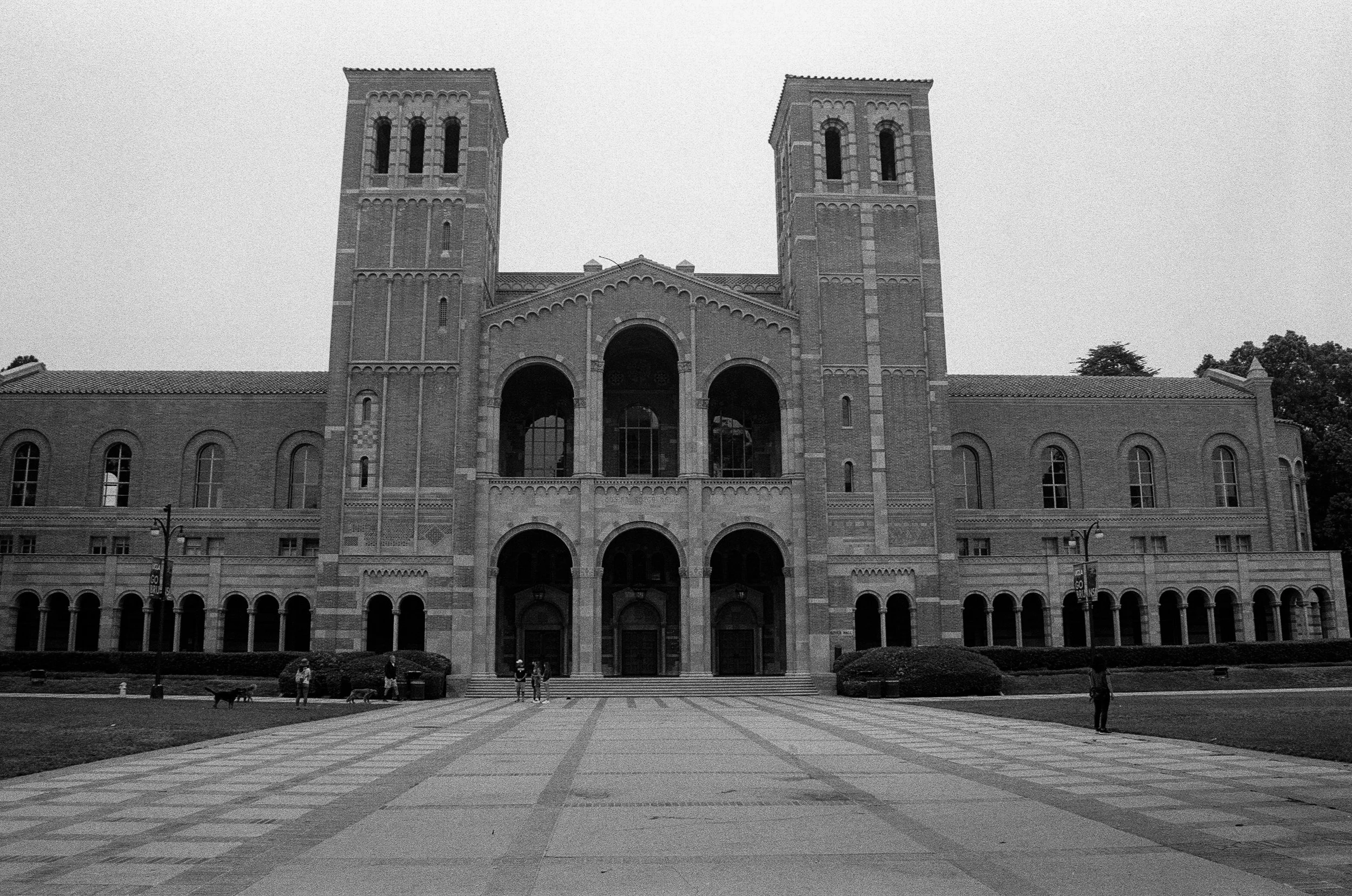 A large brick building with two tall towers and gothic architectural details, fronted by a paved open area with a few people walking and trees in the background.