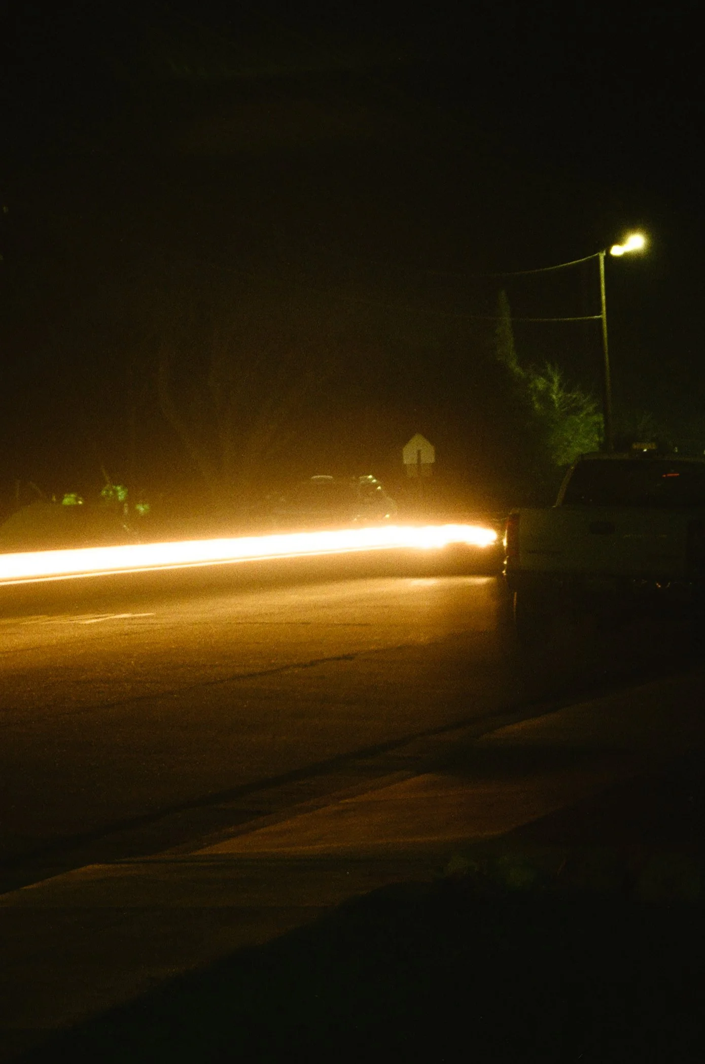 Nighttime scene of a dark street with a car in motion, illuminated by streetlights and headlights, with a visible power line and a parking lot with a few parked cars.