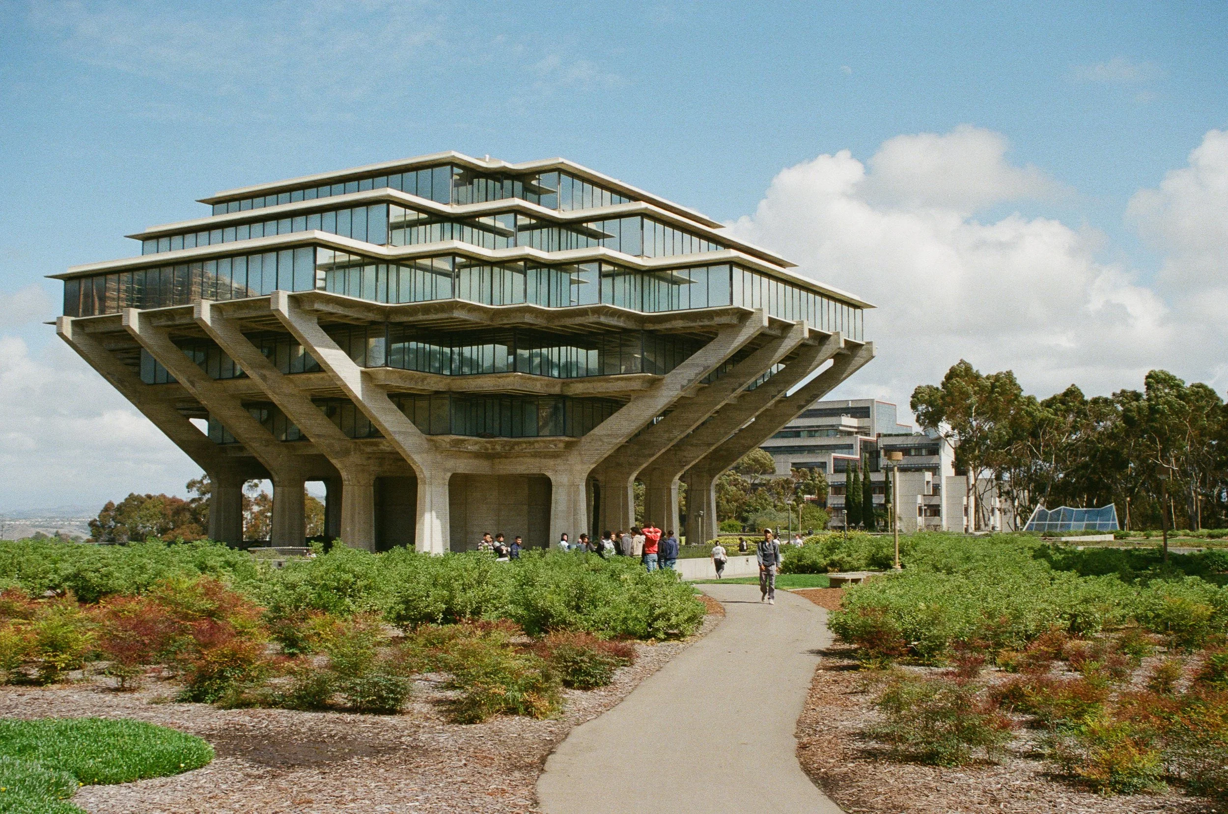 Modern building with multiple tiered floors featuring large glass windows, supported by concrete columns, situated in a park with people walking on a paved path and landscaped greenery
