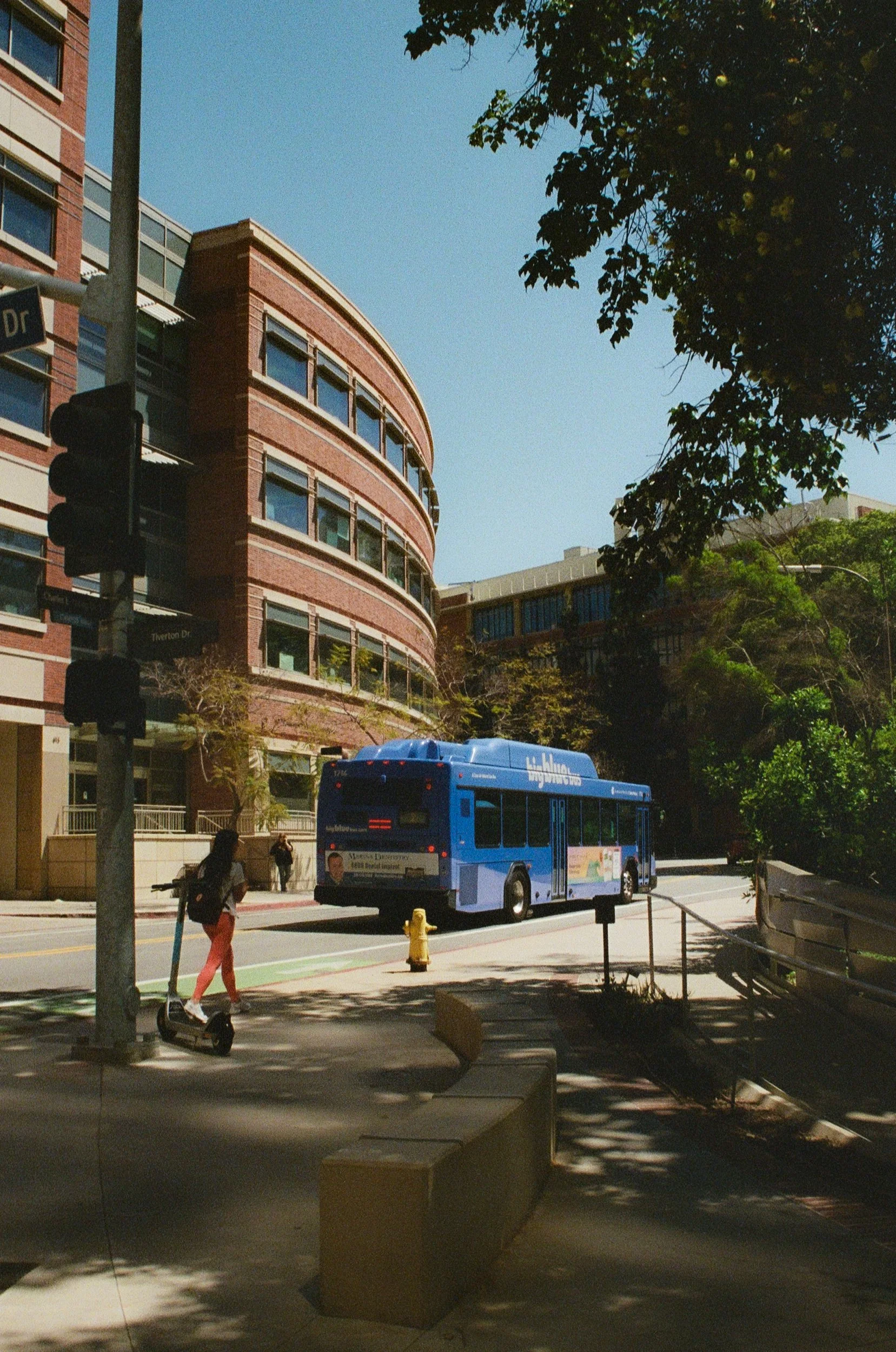 A city street scene with a blue bus, pedestrians, a traffic light, and a rounded brick building with many windows under a clear blue sky.