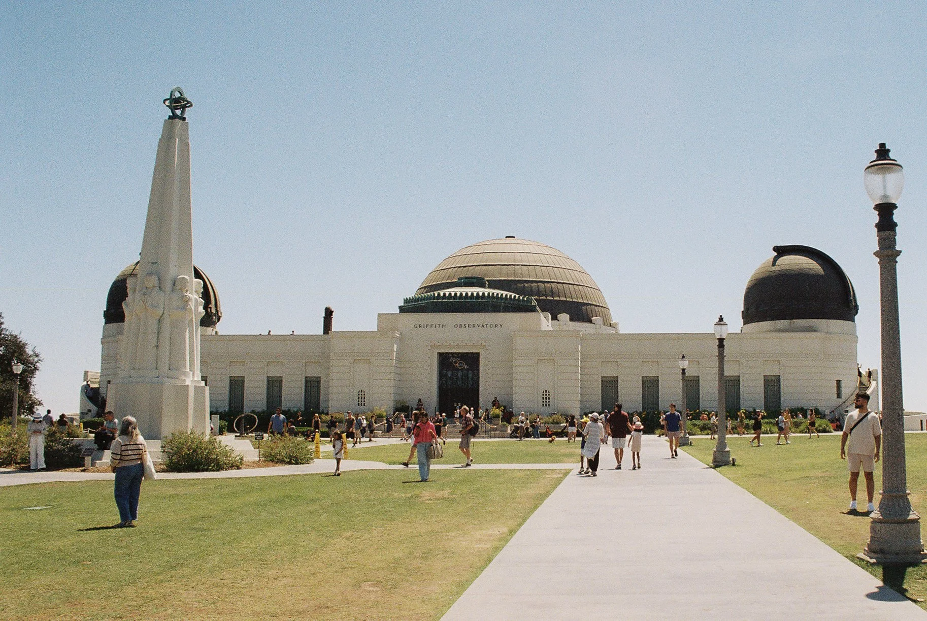 The Griffith Observatory in Los Angeles, California, with people walking on the grounds and a clear blue sky above.