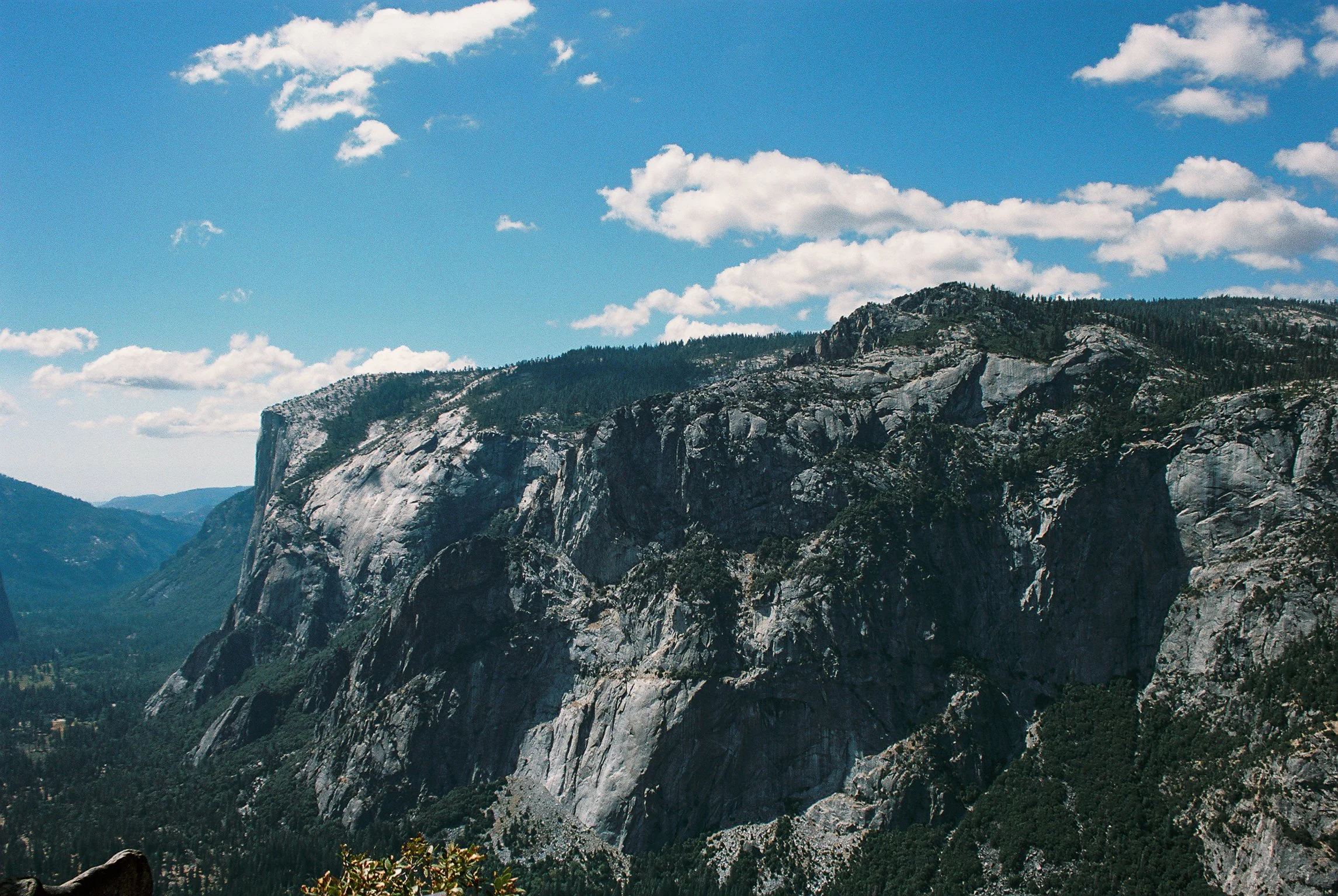 A mountain landscape with steep rocky cliffs and green forested slopes under a blue sky with scattered white clouds.