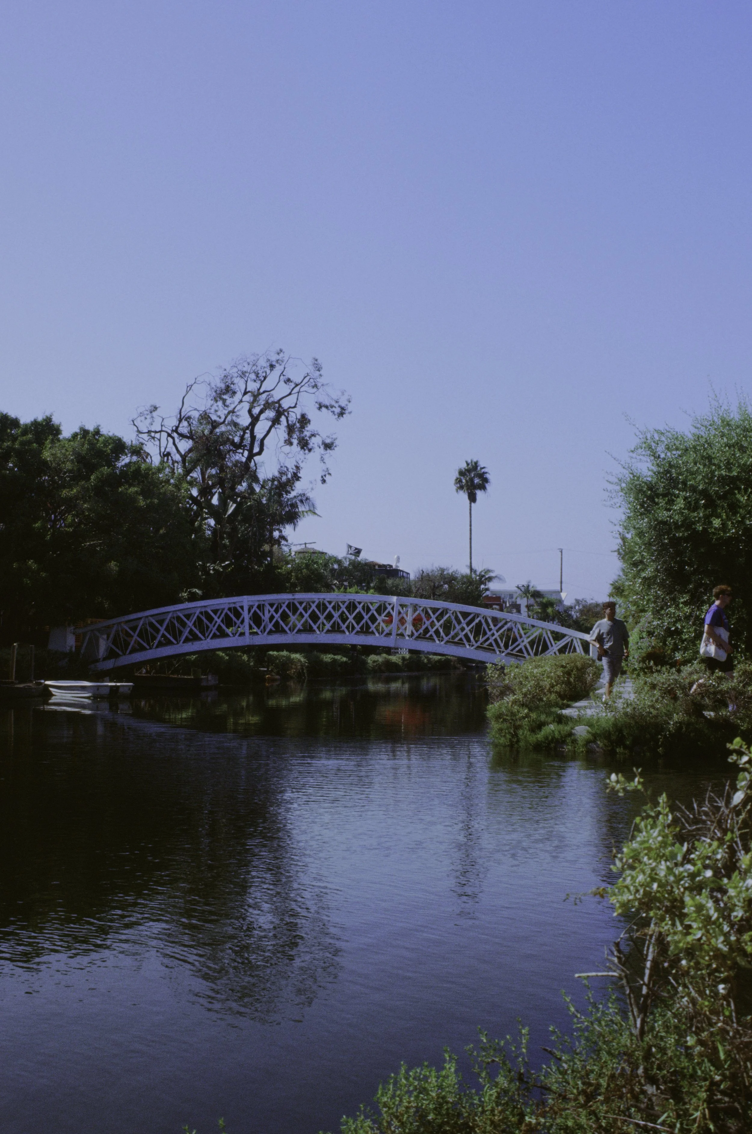 A scenic view of a small river with lush green trees on the banks, a curved pedestrian bridge over the water, and a clear blue sky overhead. Two people are walking along the path near the river.