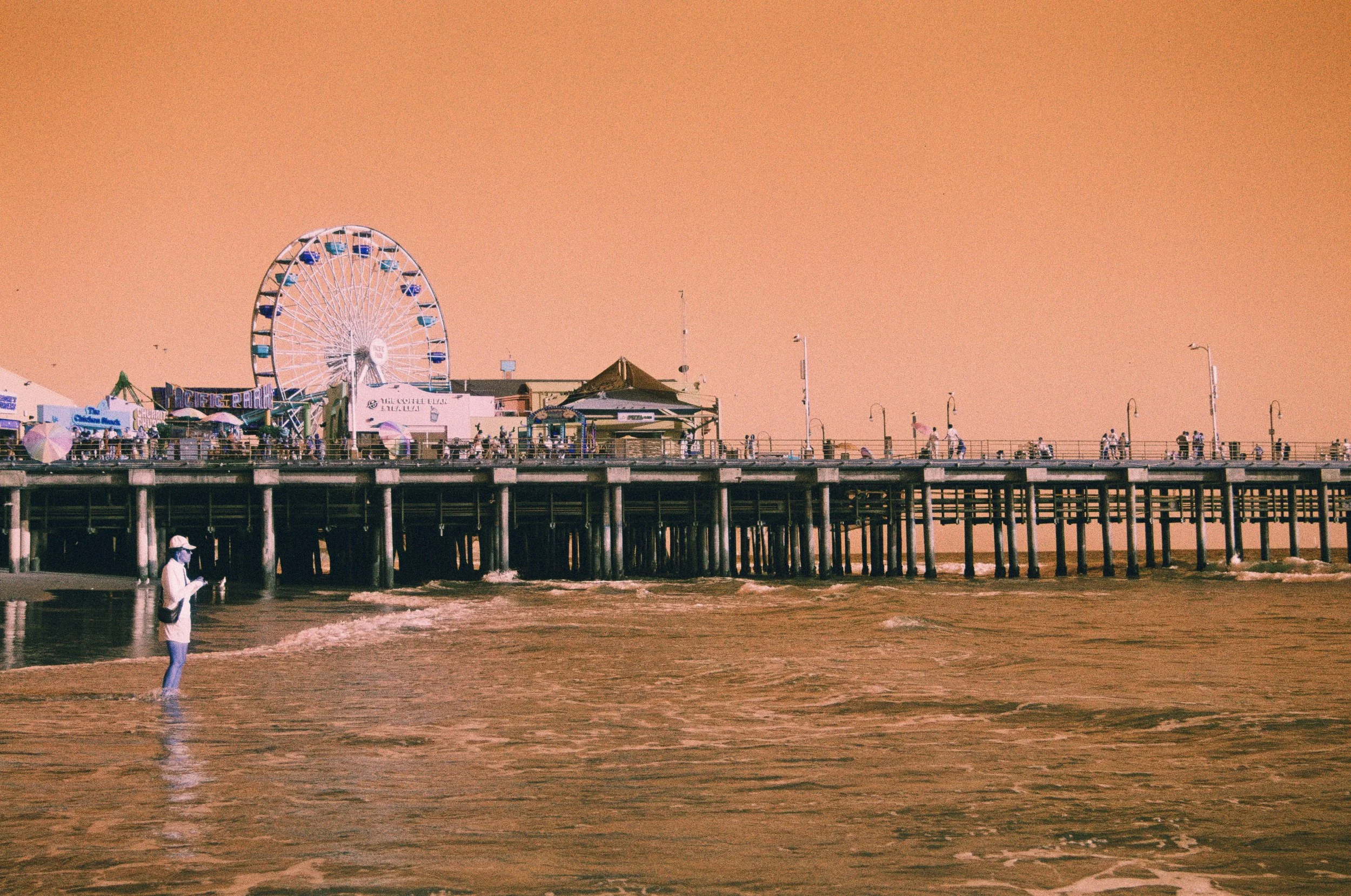 People walking and standing on a pier over the ocean with an amusement park and a Ferris wheel in the background, under a pinkish-orange sky during sunset.