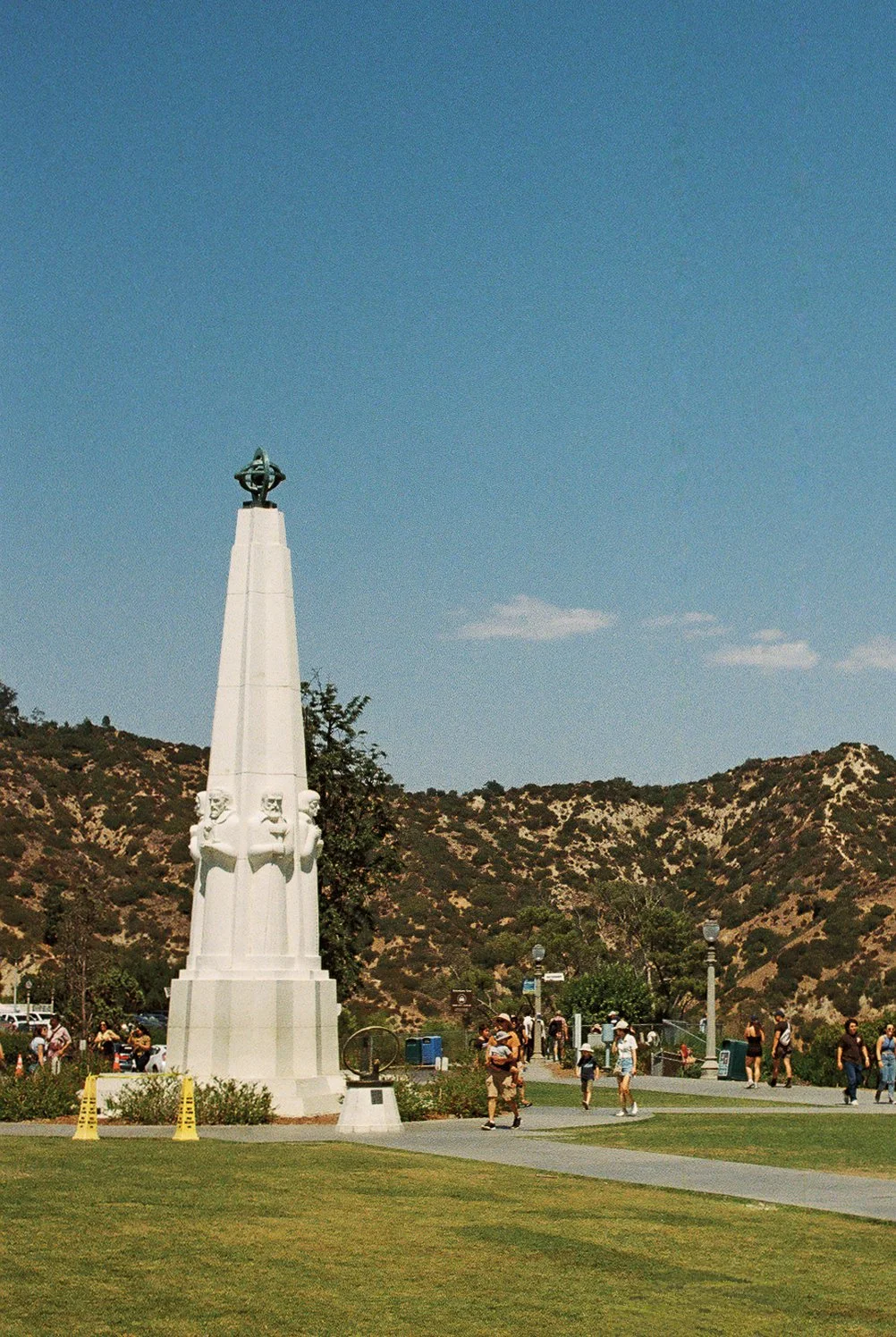 A park with a white monument and multiple people walking on pathways, with a hilly landscape in the background and a blue sky overhead.