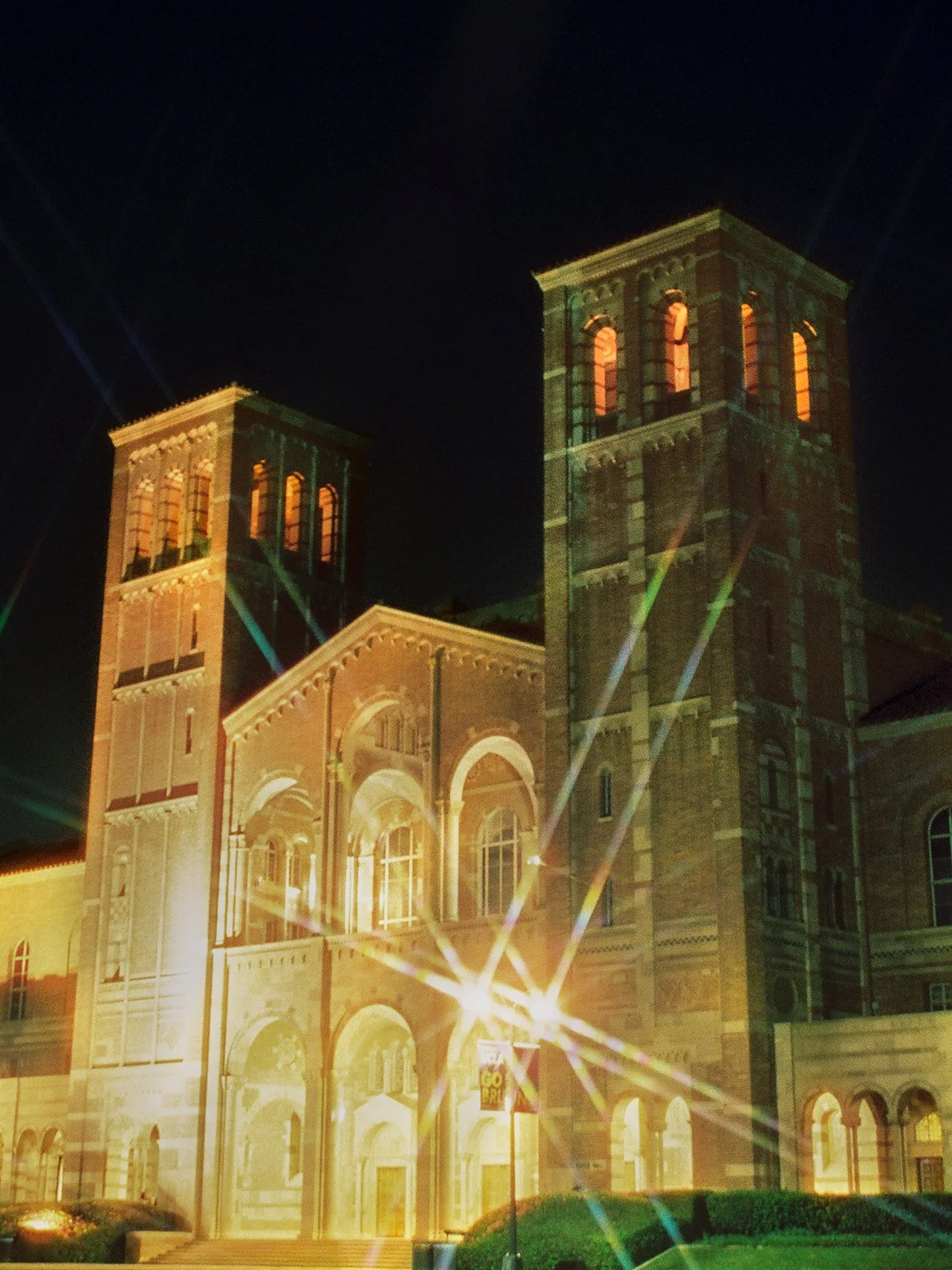Nighttime view of a large, multi-story brick building with illuminated windows, featuring towers with arched windows, bright exterior lighting, and a light display with beams projected from the ground.