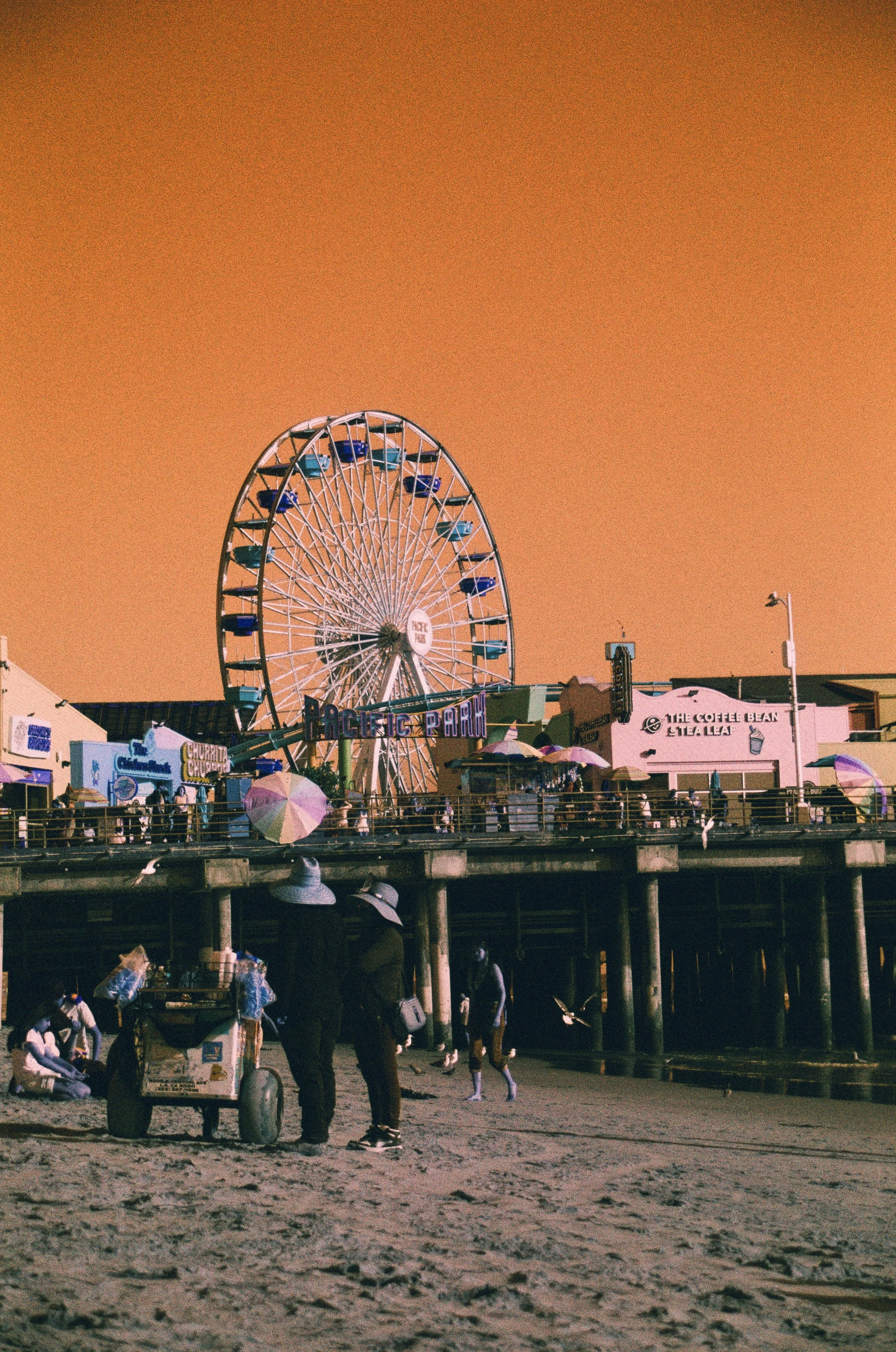 People walking along the beach promenade at sunset, with a Ferris wheel and various shops and food stands in the background.