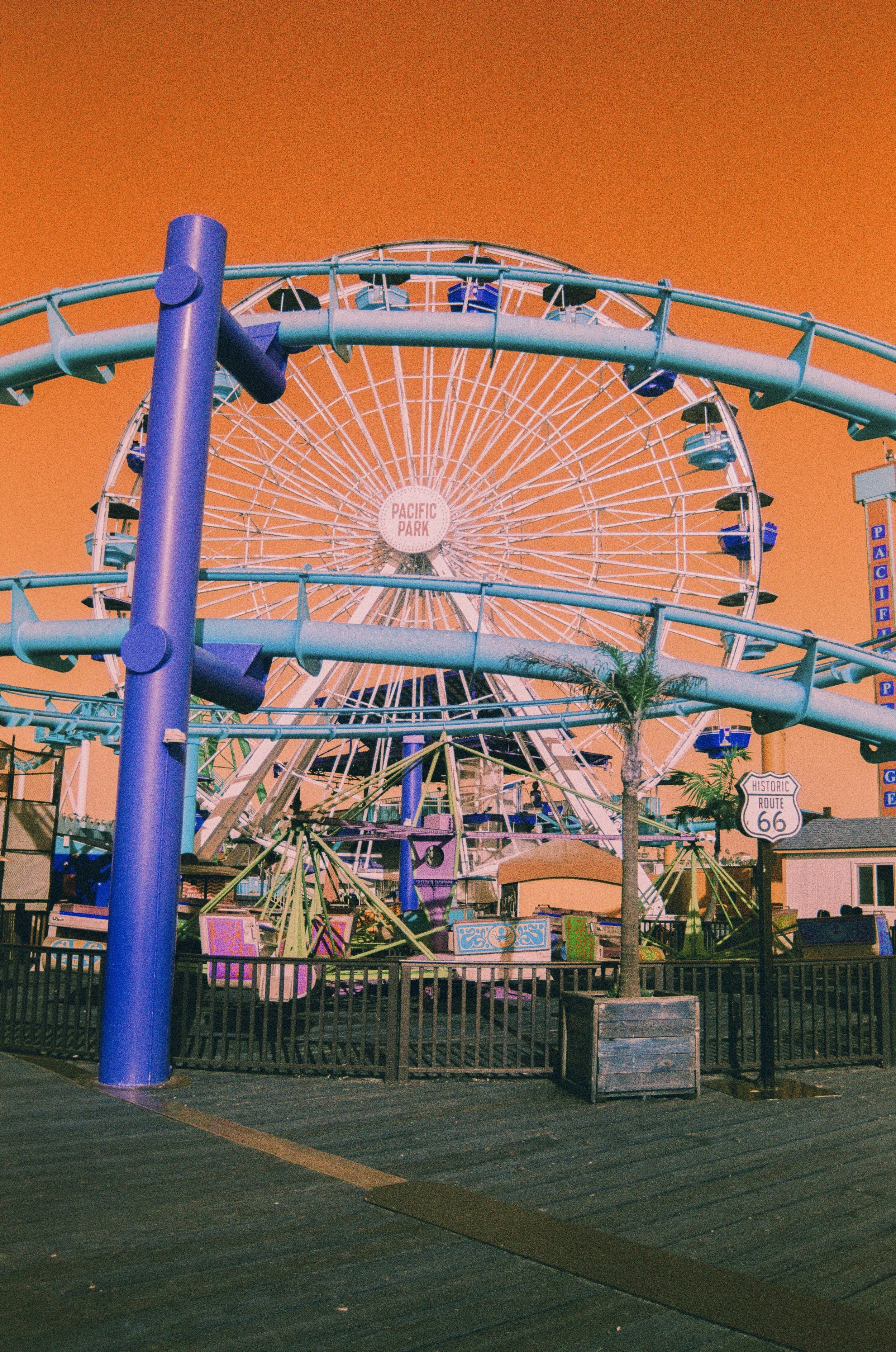 A large Ferris wheel at Pacific Park amusement park against an orange sky, with a roller coaster track in the foreground, palm trees, and a sign that says Route 66.