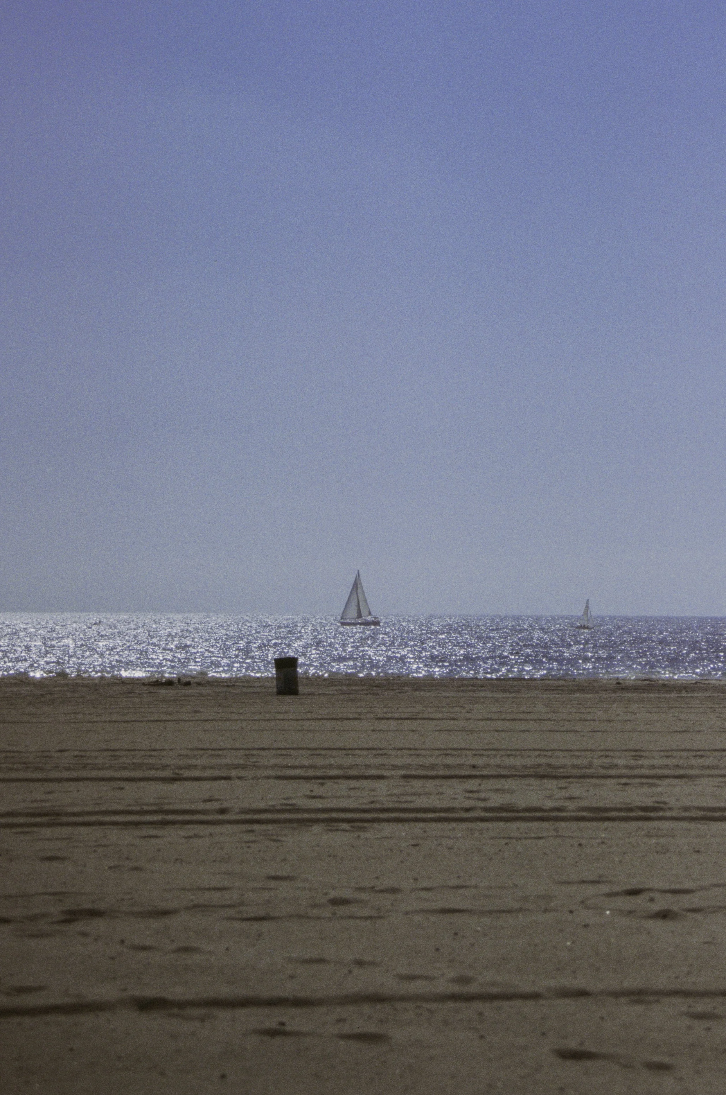 A beach scene with a sandy shoreline, a trash can, and two sailboats on the water under a clear blue sky.