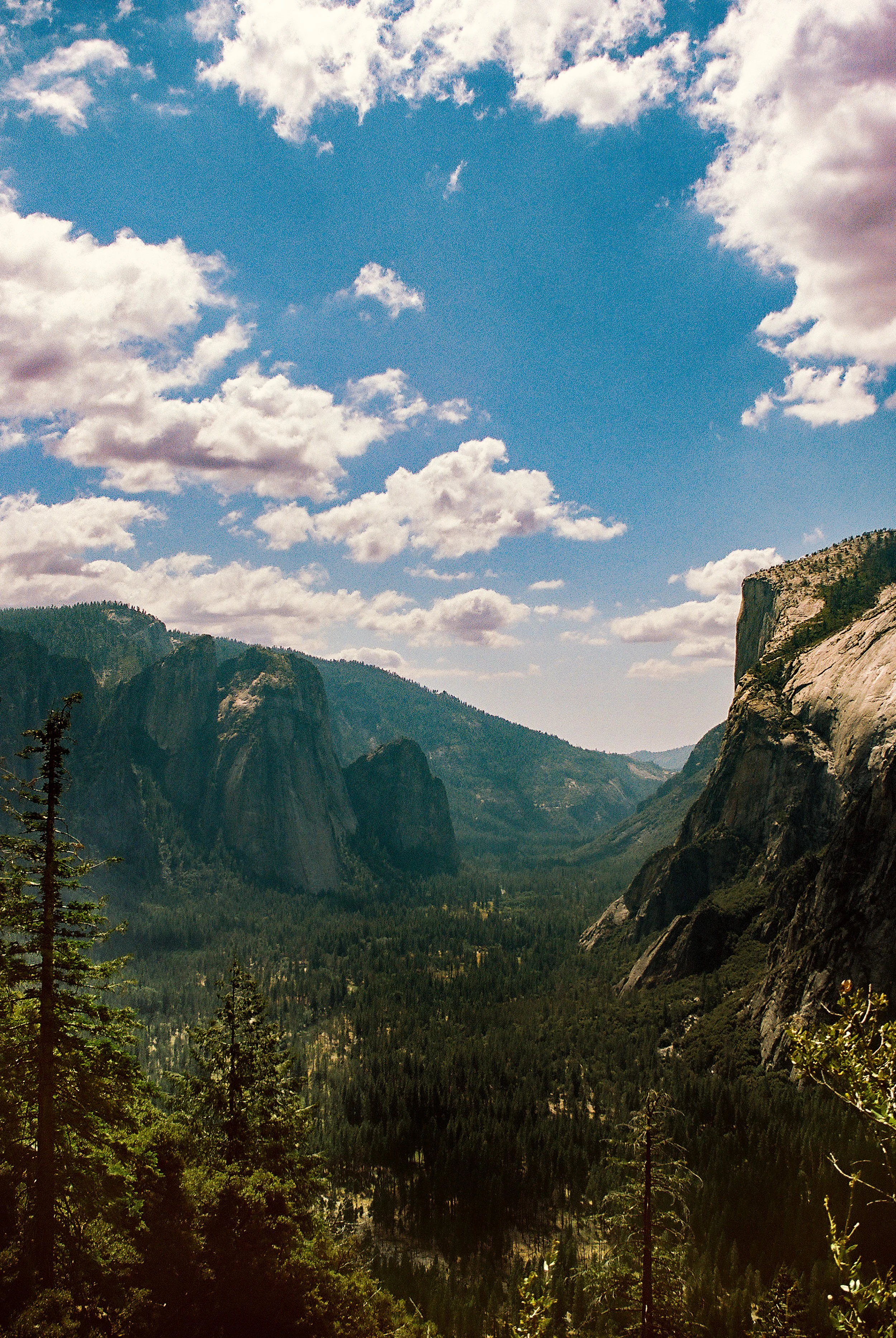 A scenic view of a mountainous landscape with tall cliffs on the right and forested valley below, under a partly cloudy sky with patches of blue.