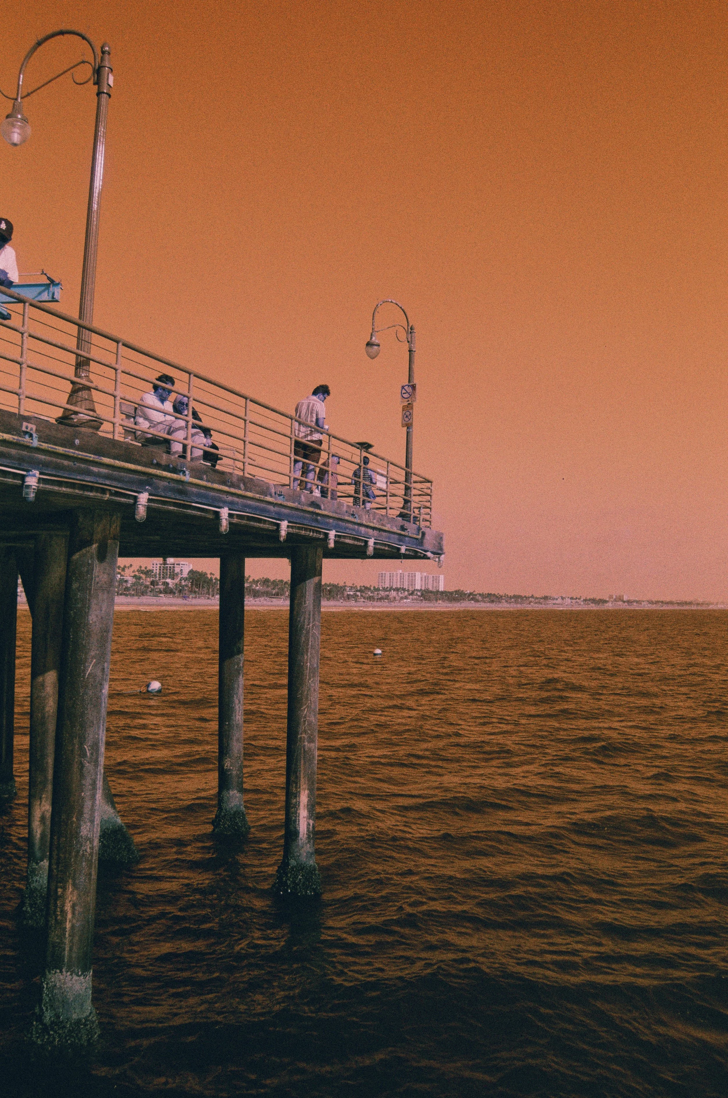 Pier extending over water at sunset or dusk, with several people sitting and standing on it, and a city skyline in the background.