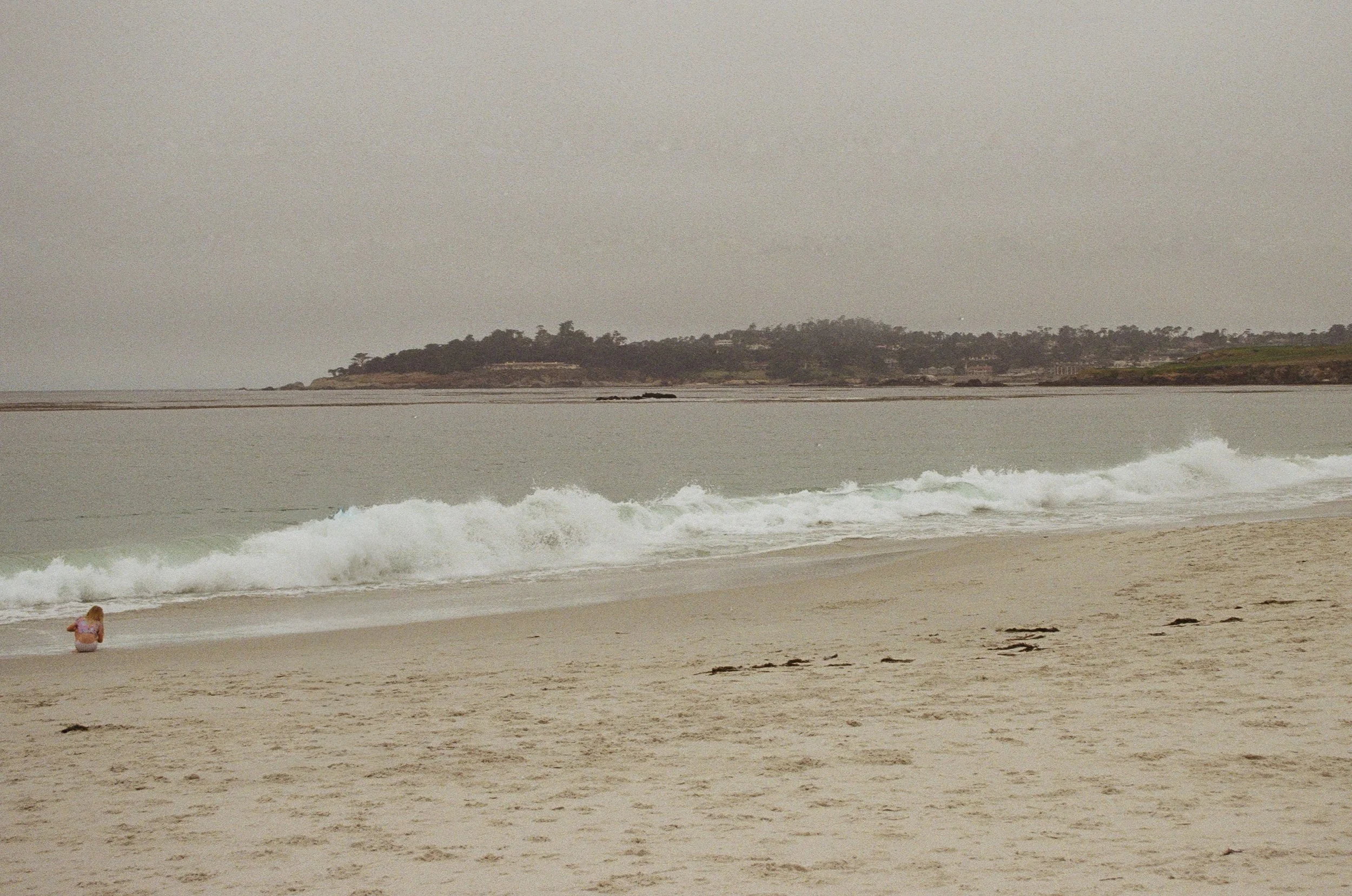 A person sitting on the sandy beach near the shoreline, with ocean waves and a distant landmass covered in trees and buildings under a cloudy sky.