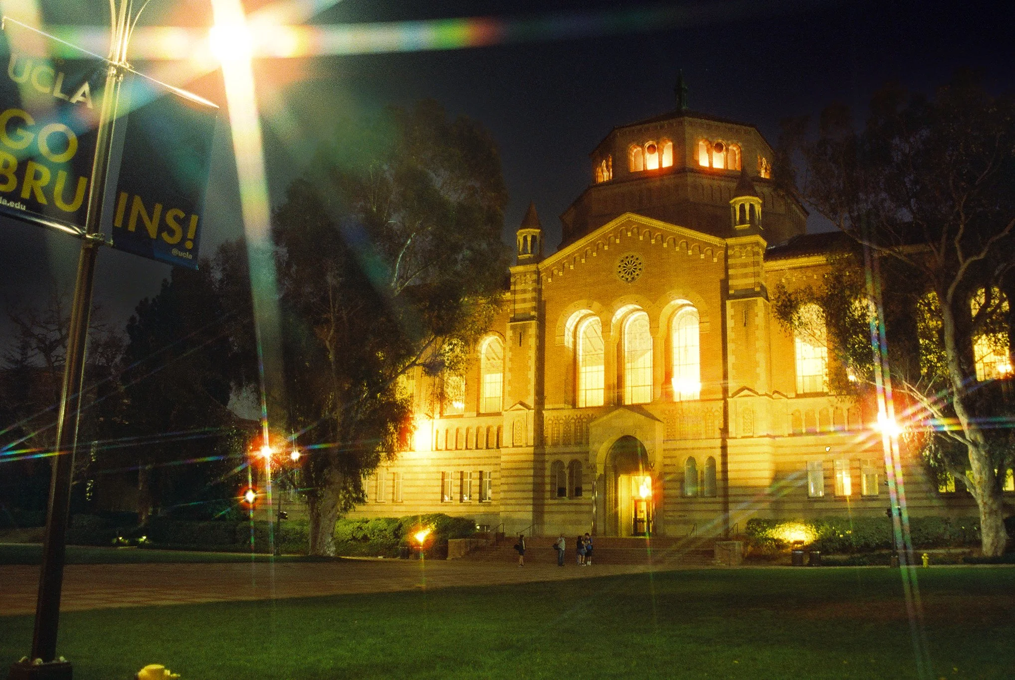 Night view of a historic building with illuminated windows and a dome, surrounded by trees and a park with people walking. There is a sign on the left that reads 'UCLA' and 'GO BRU!'. Flare effects from nearby streetlights create rainbow-like light s