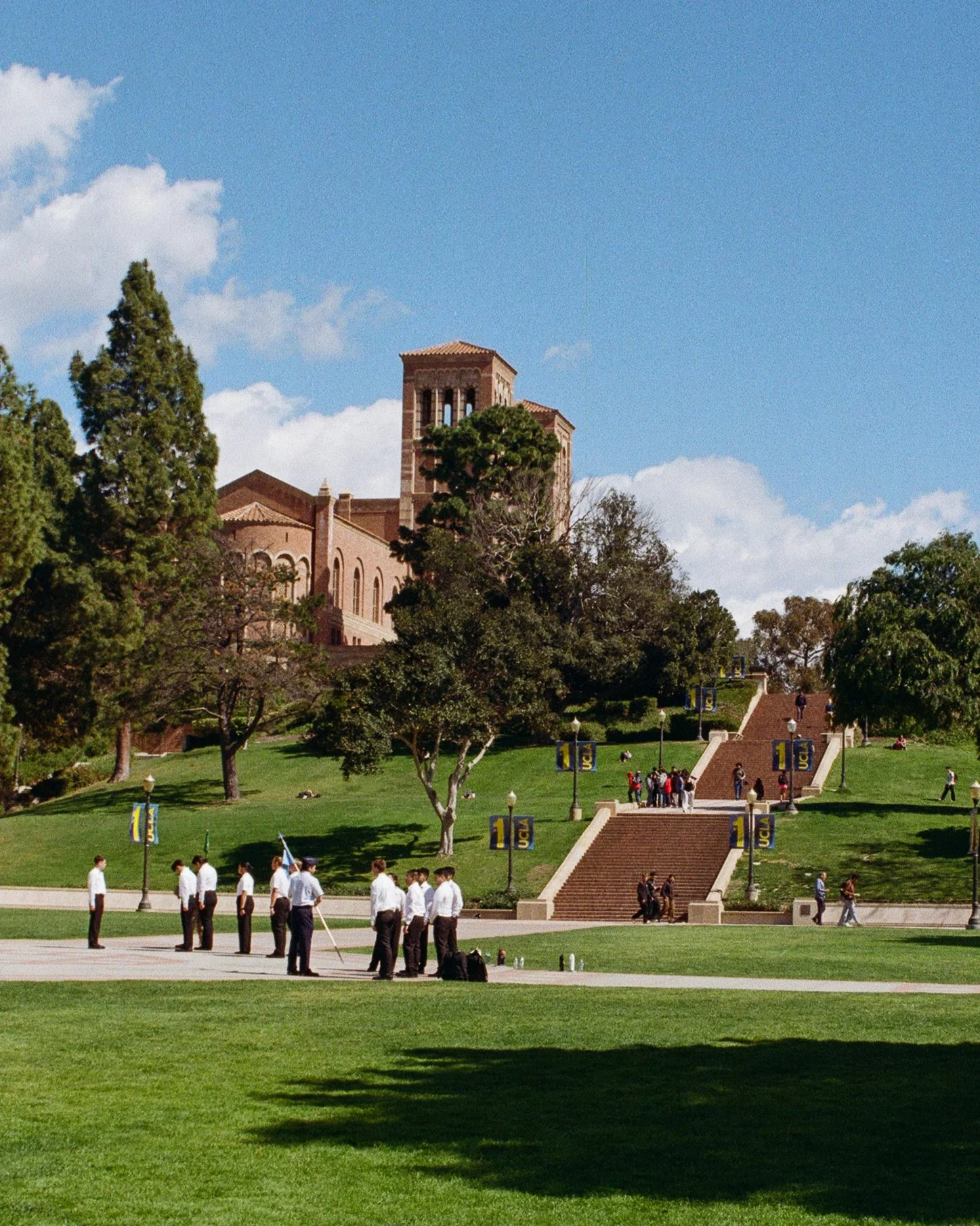 UCLA campus with students gathered on green lawn, stairs leading up to university buildings, and a clock tower in the background under a partly cloudy sky.