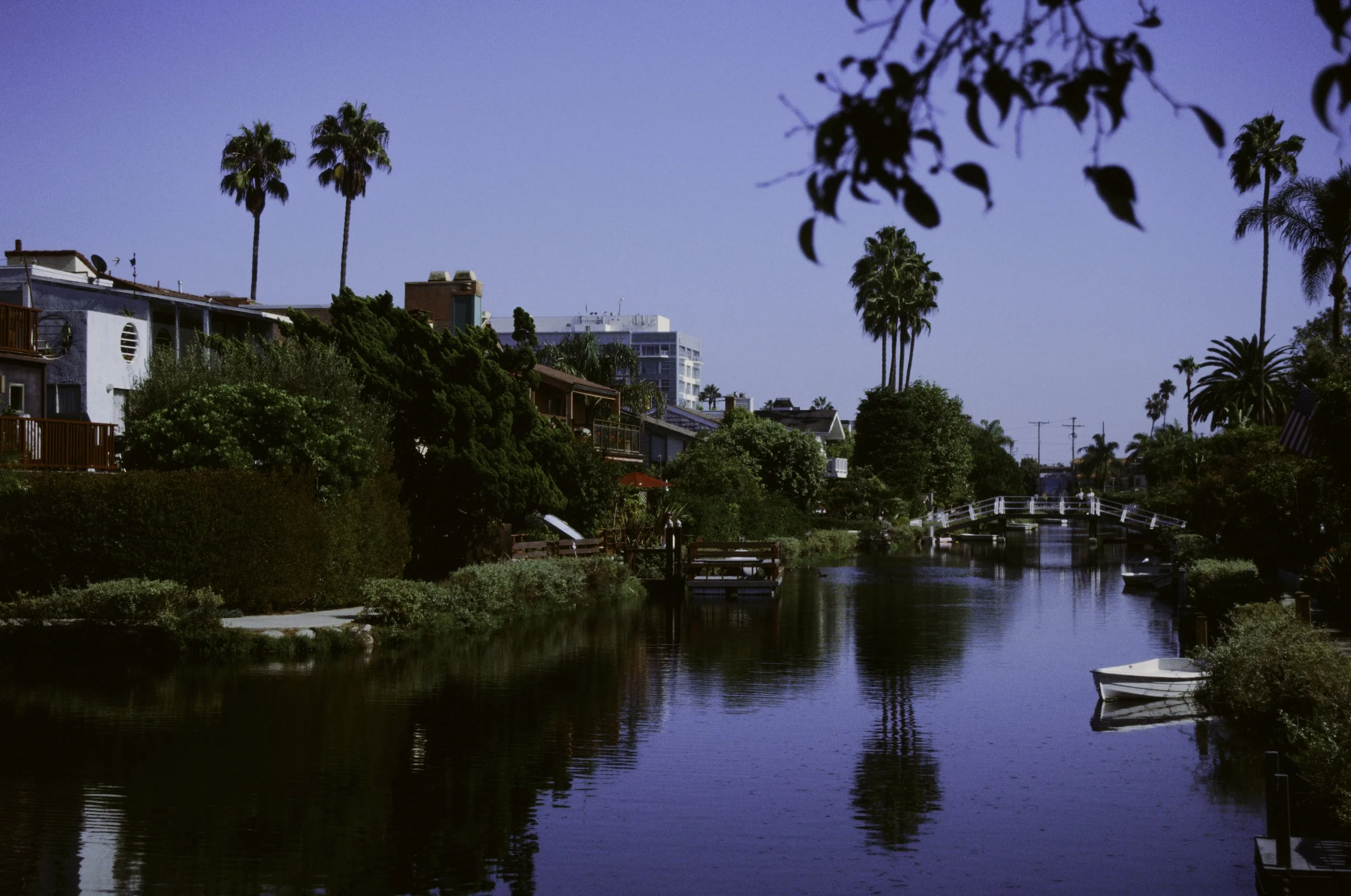A scenic view of a small canal lined with houses, trees, and palm trees, with a bridge over the water and a boat docked at the edge, under a clear blue sky.