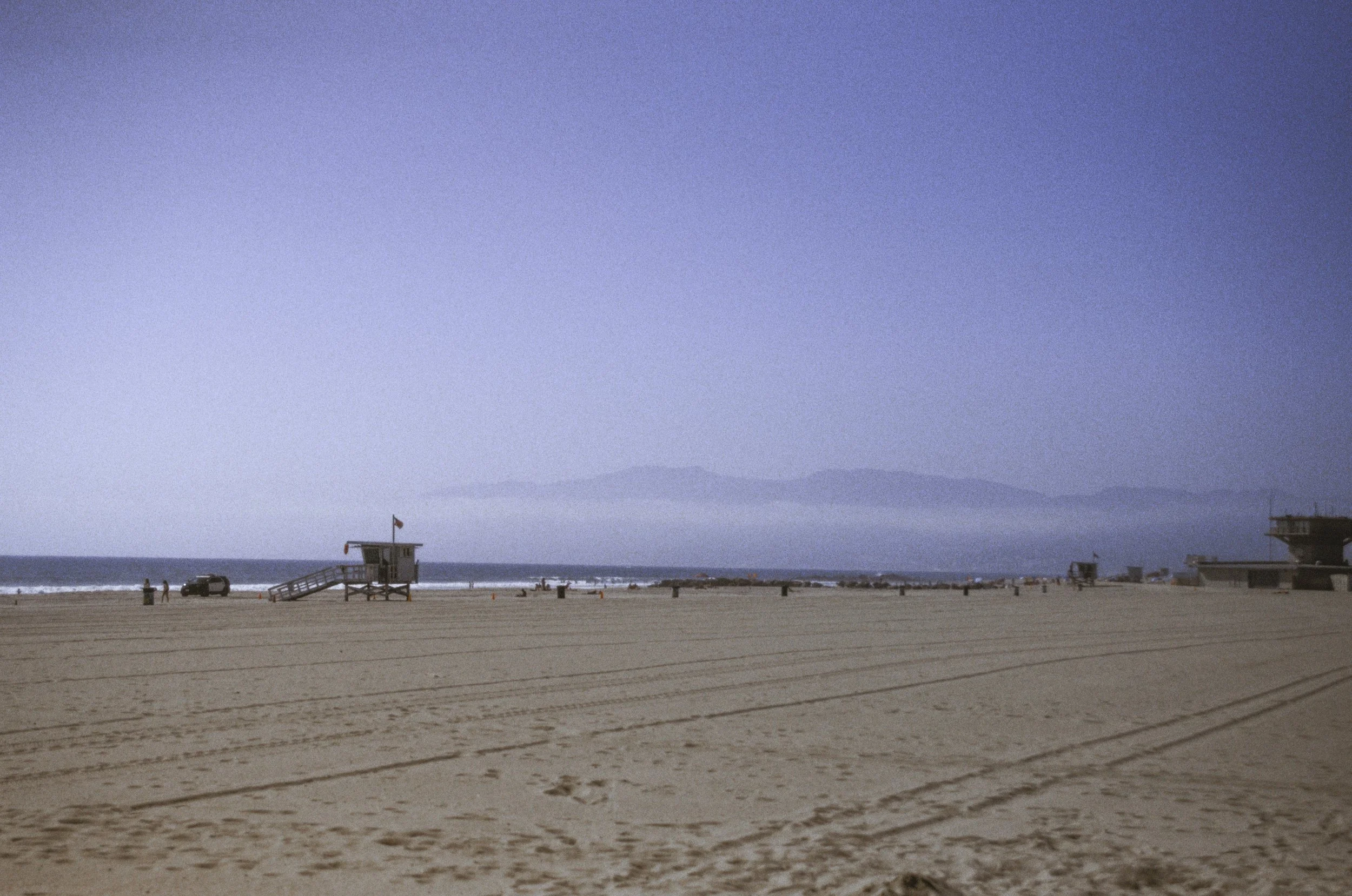 A sandy beach with a lifeguard tower, vehicles, and a few people near the shoreline, with ocean waves and mountains in the background under a blue sky.