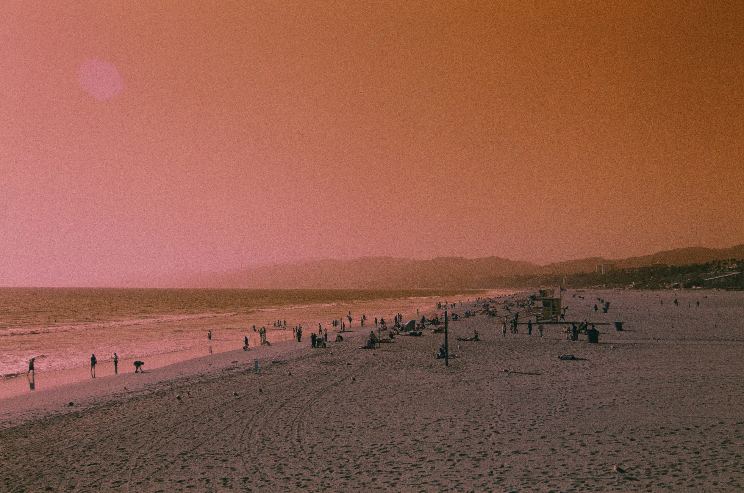 A beach scene during sunset with people walking and sitting along the shore, lifeguard towers, and a distant mountain range under an orange-pink sky.