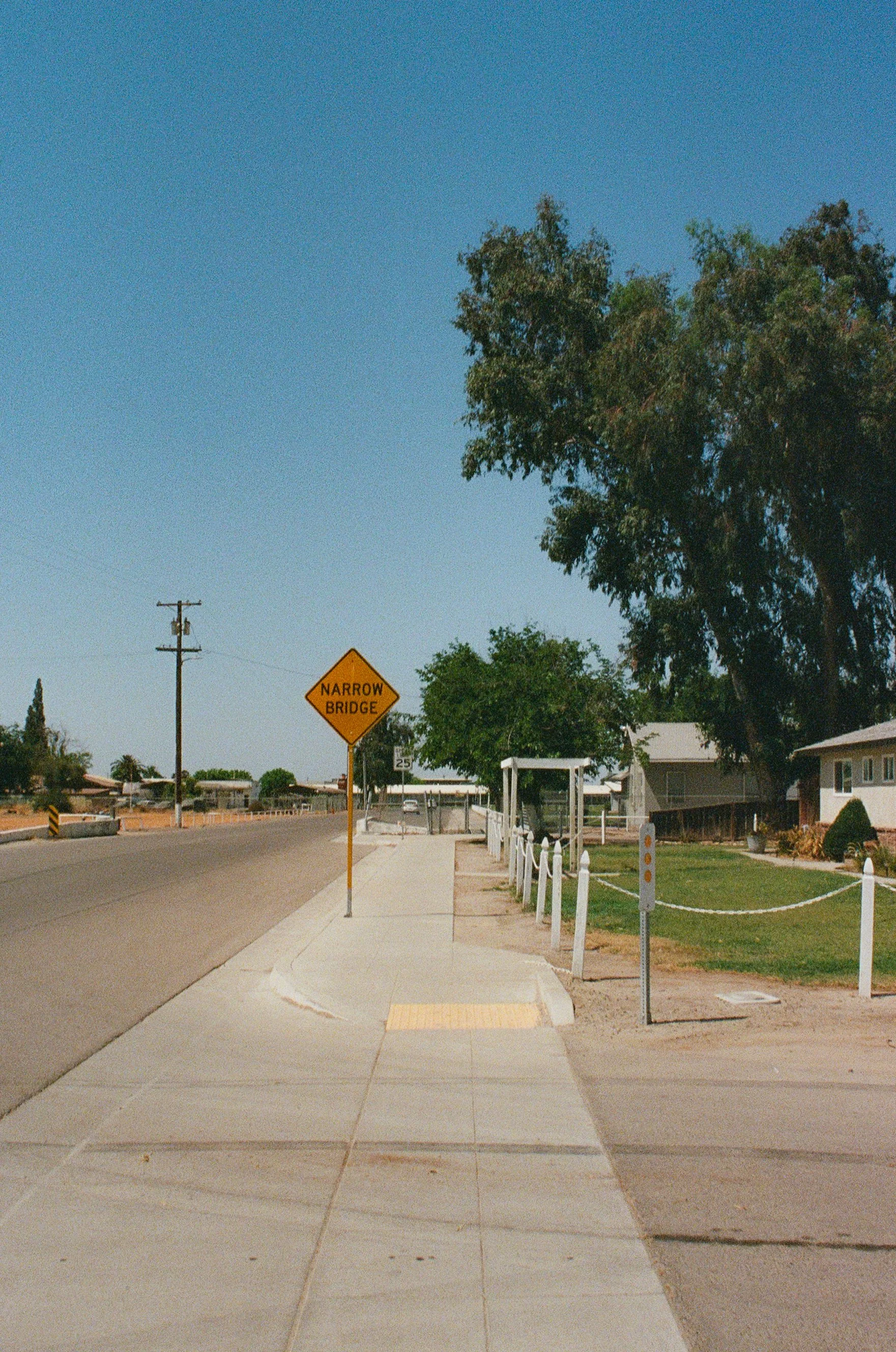 A sidewalk with a yellow tactile paving strip for the visually impaired, a street sign that reads 'Narrow Bridge,' and residential houses with lawns and trees under a clear blue sky.