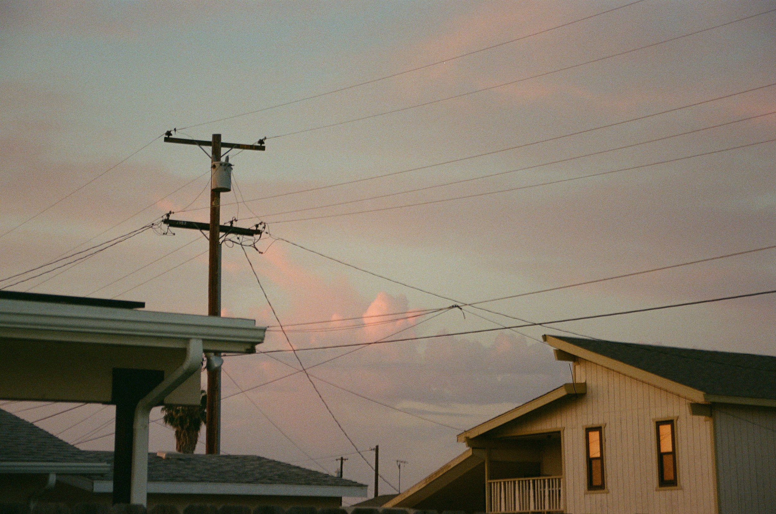 Residential neighborhood with rooftops, a utility pole with multiple power lines, and a pastel-colored sky with soft pink and orange clouds at dusk or dawn.