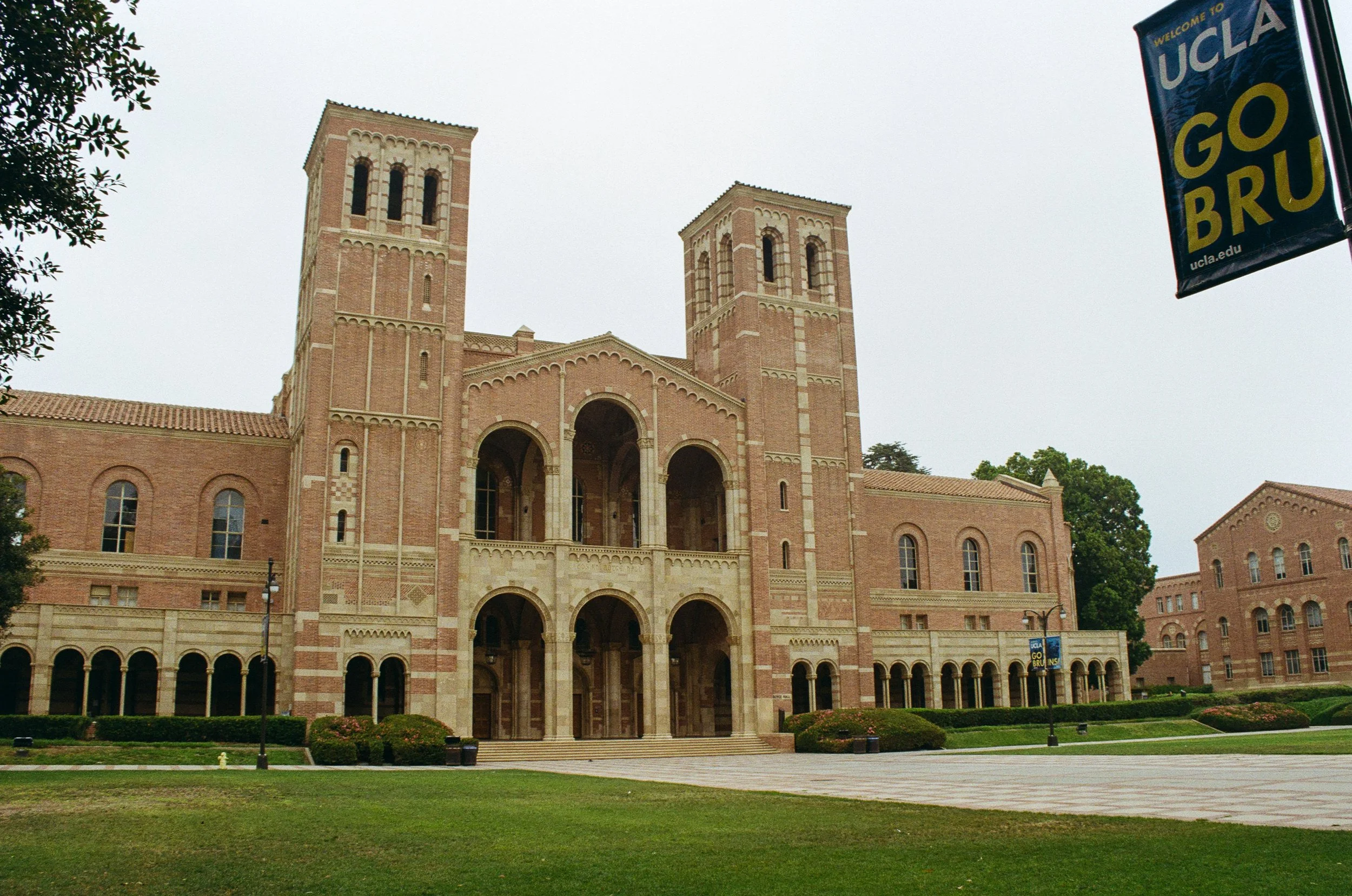 A brick university building with two tall towers, arched entrances, and a landscaped lawn in front, with a UCLA welcome banner hanging to the right.