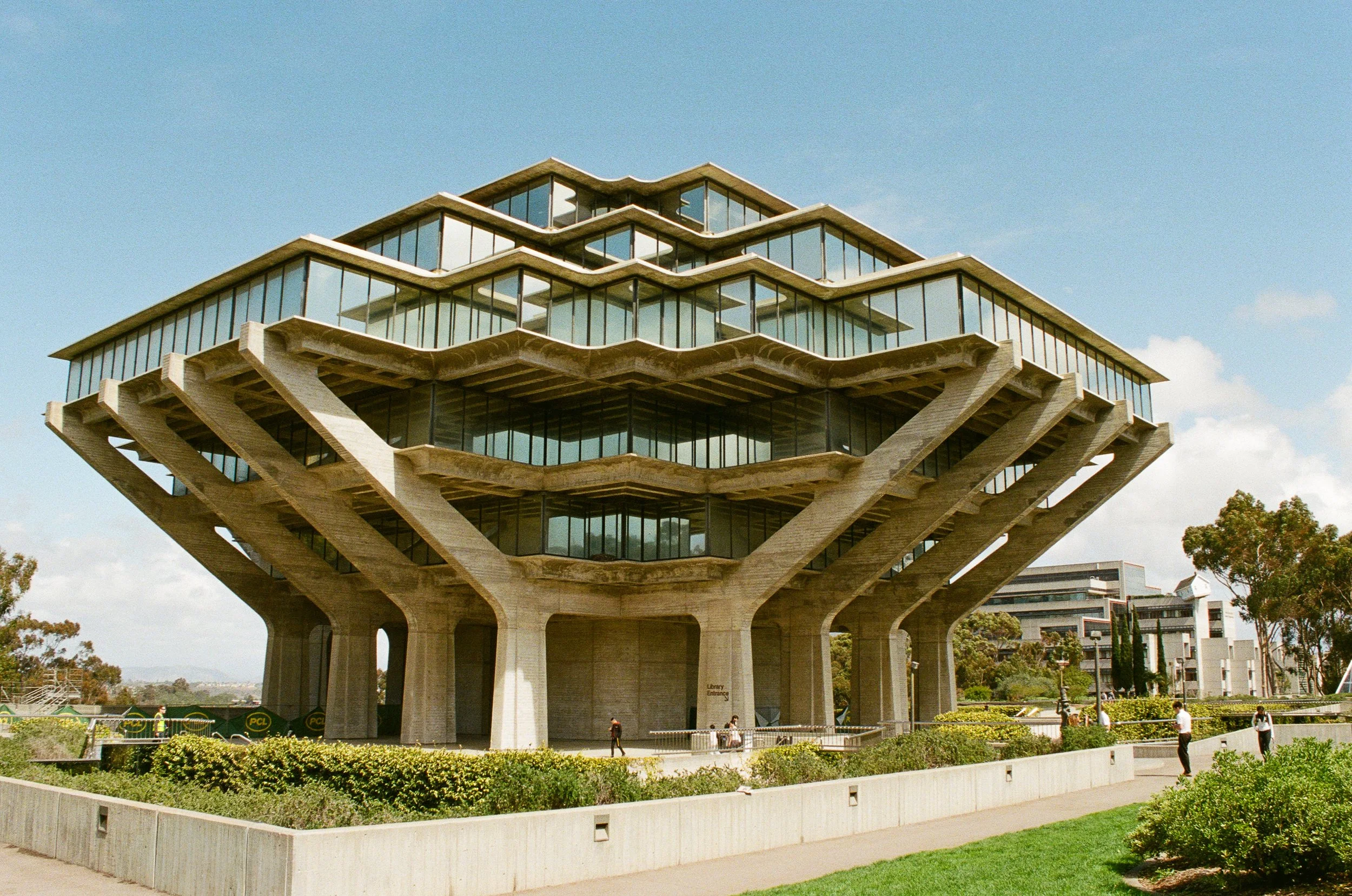 A modern multi-tiered building with large glass windows and unique architectural design, supported by concrete pillars, with people walking near it and green landscaping in the foreground, under a clear blue sky.