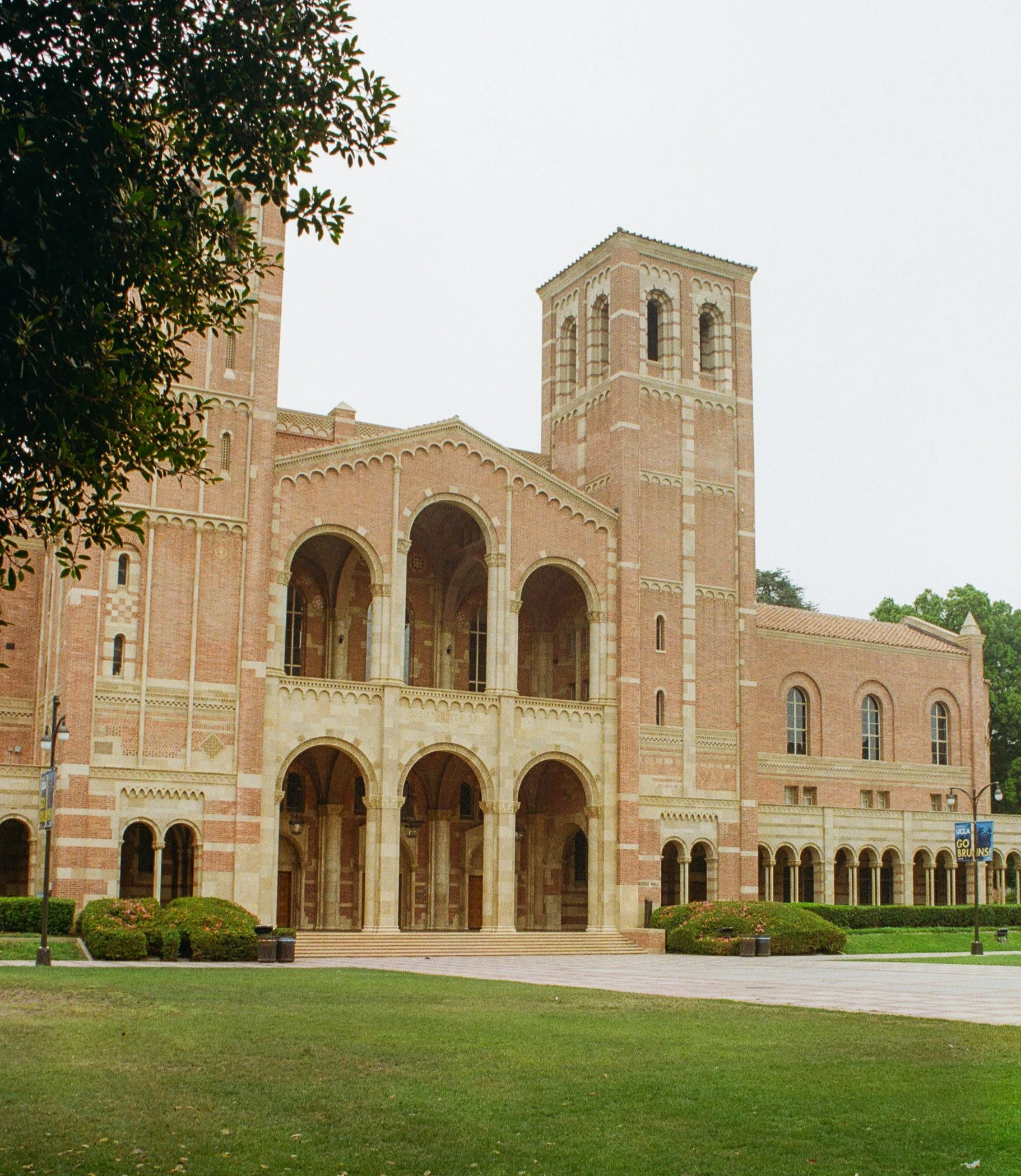 A large brick church with tall bell towers, arched windows, and a wide staircase leading to the entrance, surrounded by a well-maintained lawn and some bushes, overcast sky.