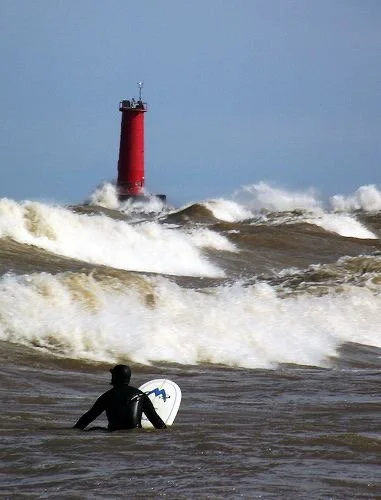 A surfer in the lake facing large waves with a red lighthouse in the background.