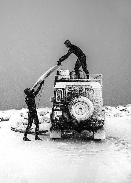 Two people in wet suits passing a surfboard on a snow-covered vehicle in a snowy landscape, with snow falling.