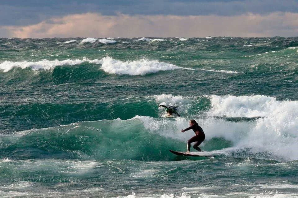 Two surfers riding on lake waves during daytime, one standing and one lying on the board, with a cloudy sky in the background.