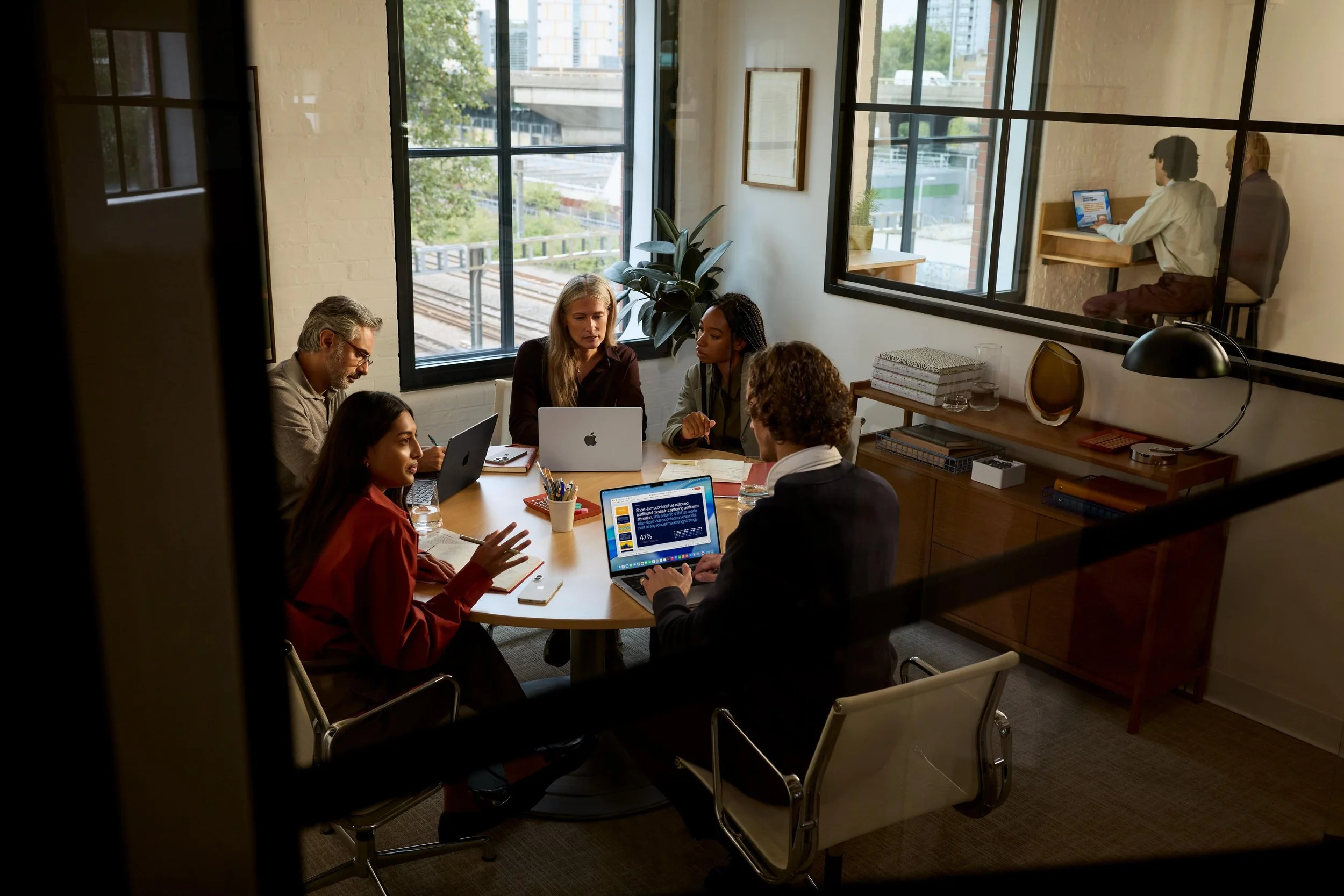 A group of six diverse professionals gathered around a conference table in a modern office, engaging in a discussion with laptops and notebooks, with a large window showing an urban cityscape in the background.