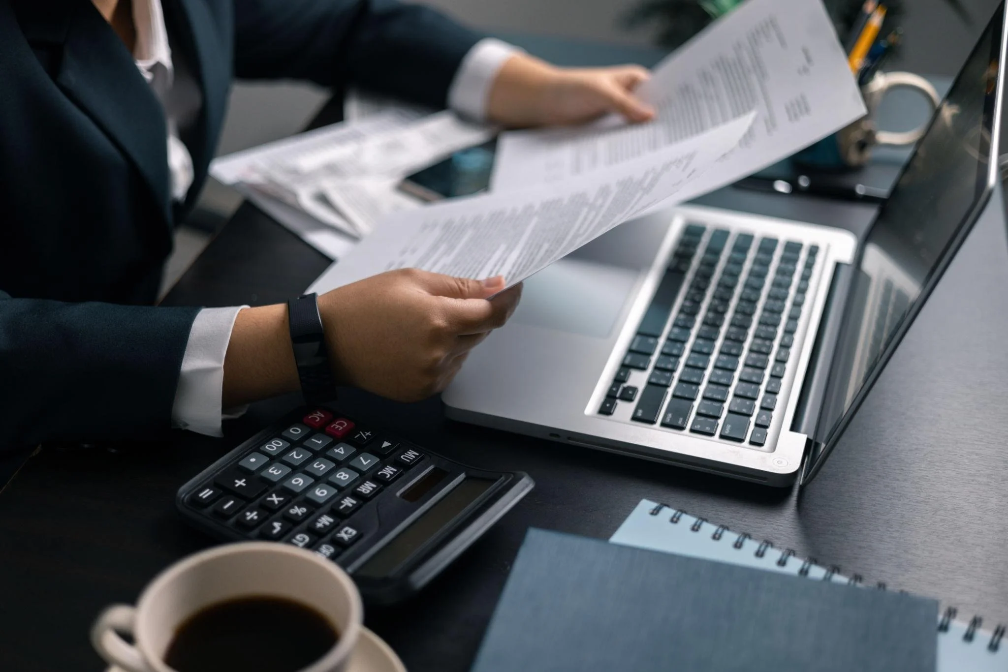 Businessperson holding a sheet of paper with financial documents, sitting at a desk with a laptop, calculator, and coffee cup.