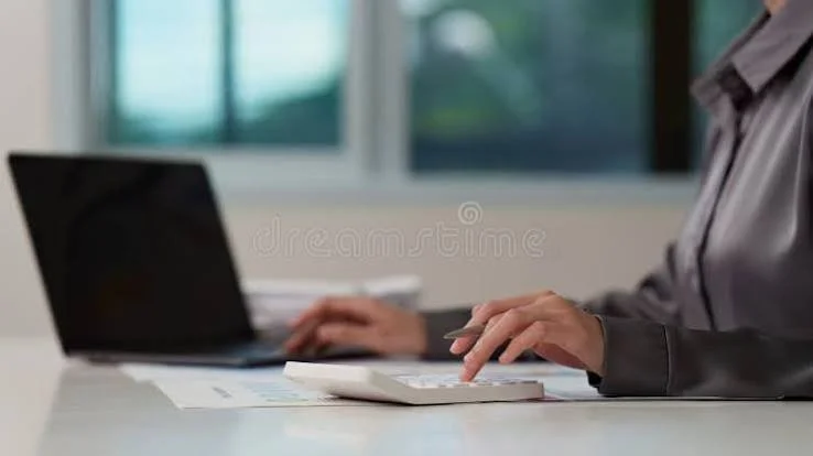 Person working at a desk with a calculator and a laptop in front of a window.