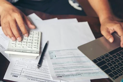 Person working at a desk with financial documents, calculator, pen, and laptop.