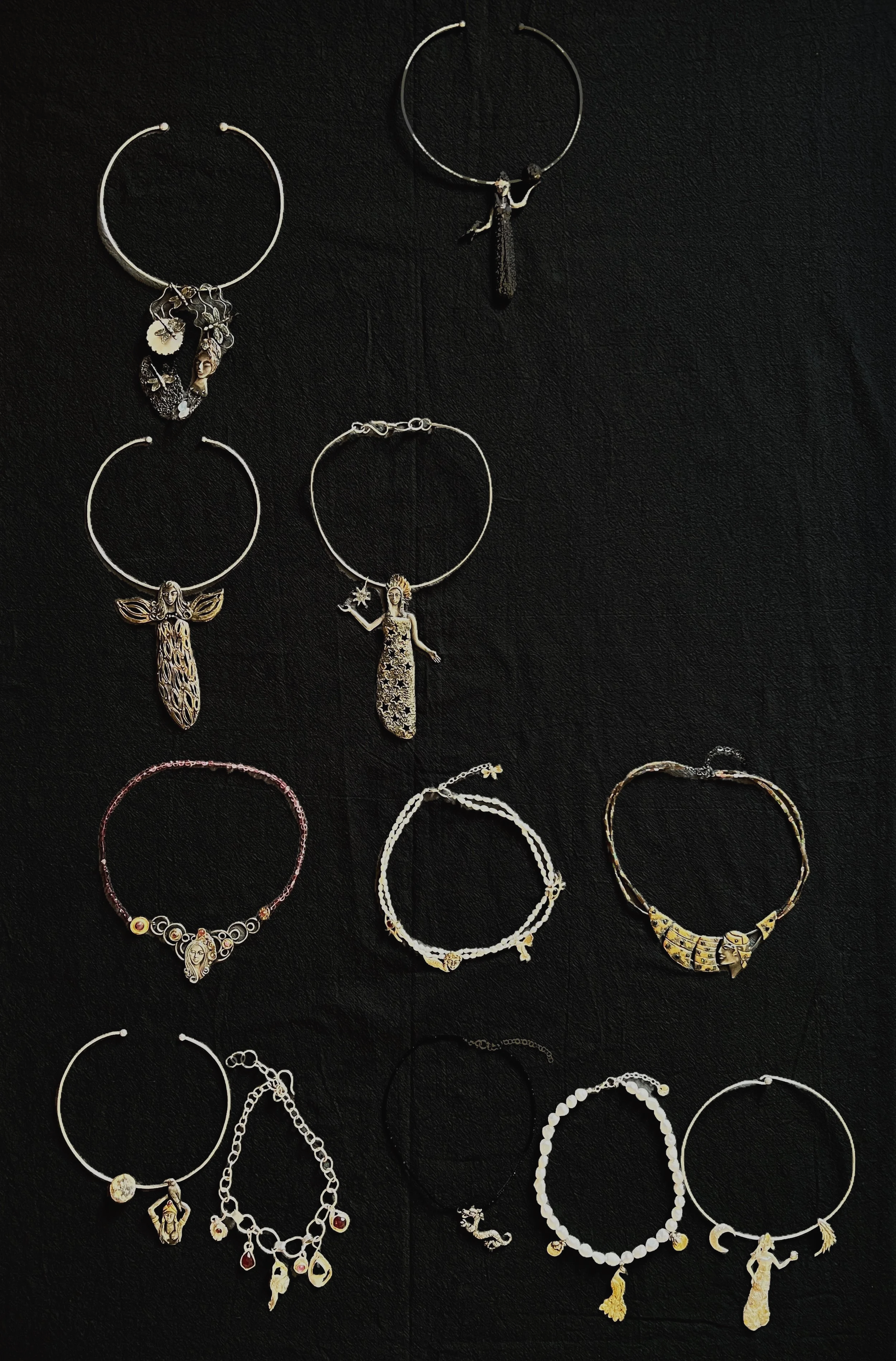 A collection of silver and gold jewelry, including necklaces and earrings, displayed on a black background. The jewelry features various charms, pendants, and decorative elements with intricate designs.