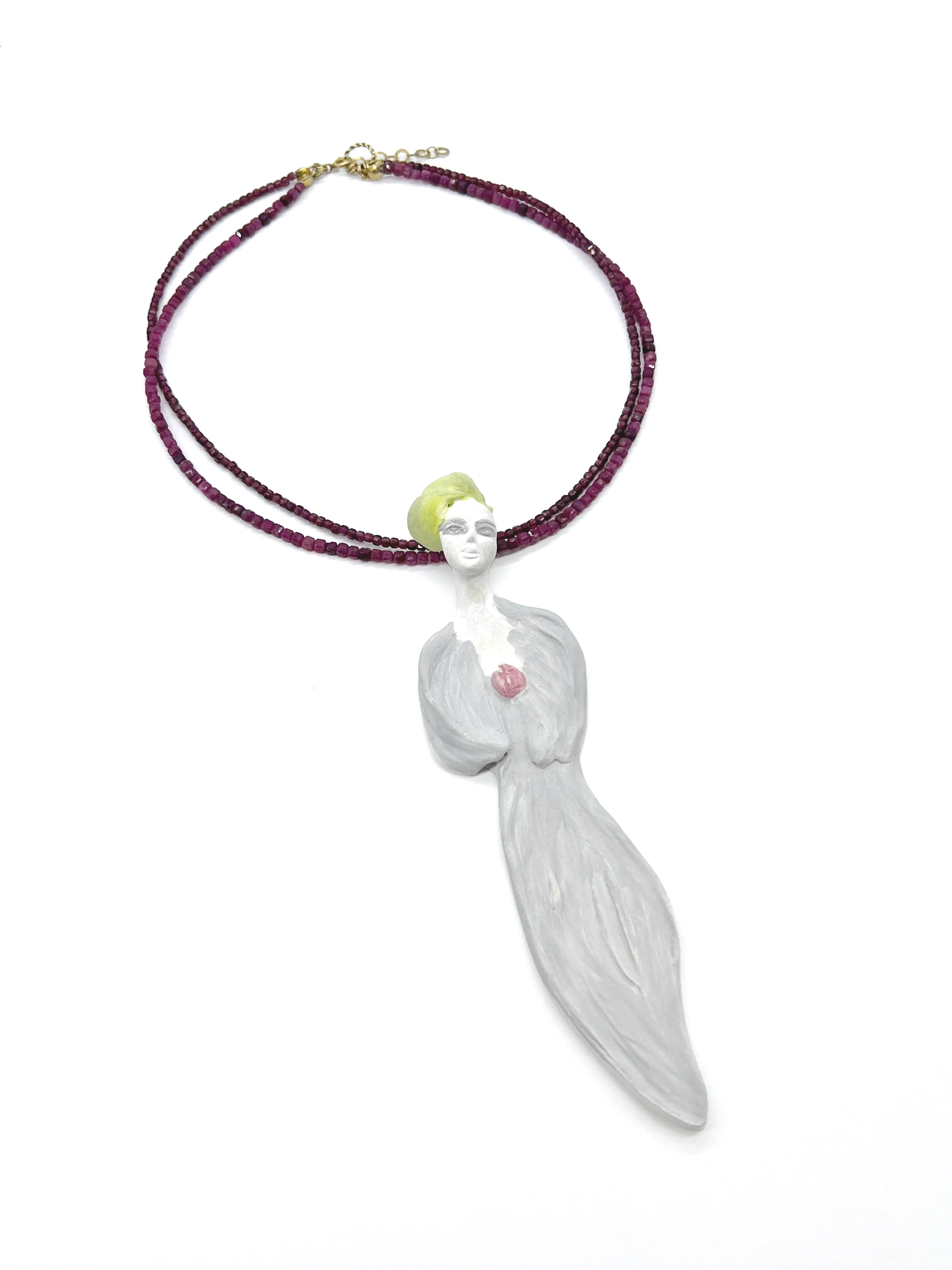 A white sculpture of a woman with short yellow hair, holding her hands over her chest with a pink heart-shaped object, placed under a double-stranded purple beaded necklace with a small figurine clasp on a white background.
