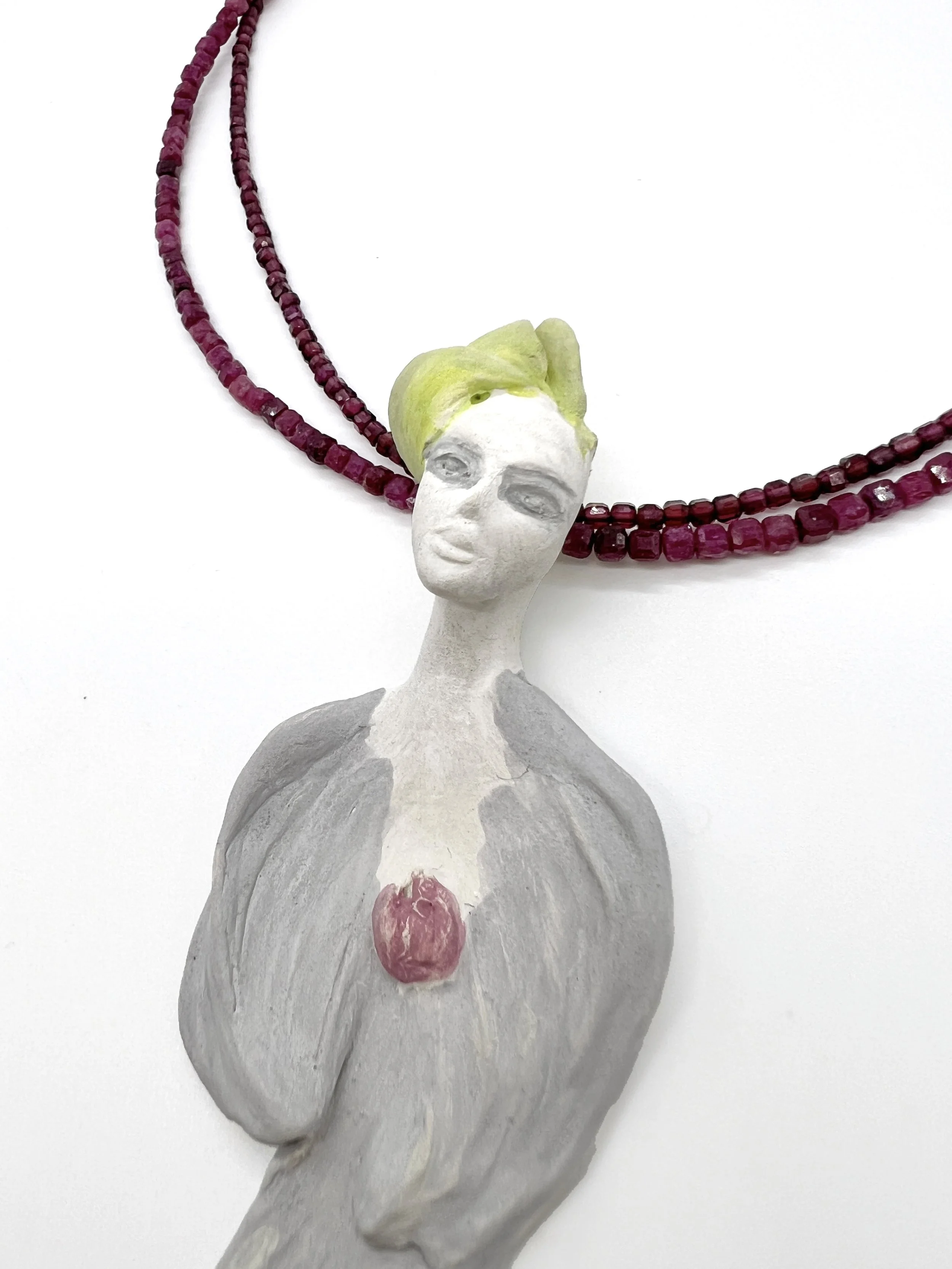 A sculpture of a female figure with painted hair, wearing a leafy crown, with a necklace made of pink beads and a pink heart-shaped pendant on her chest. The sculpture's face is painted with makeup, and she has a long neck and sloped shoulders.