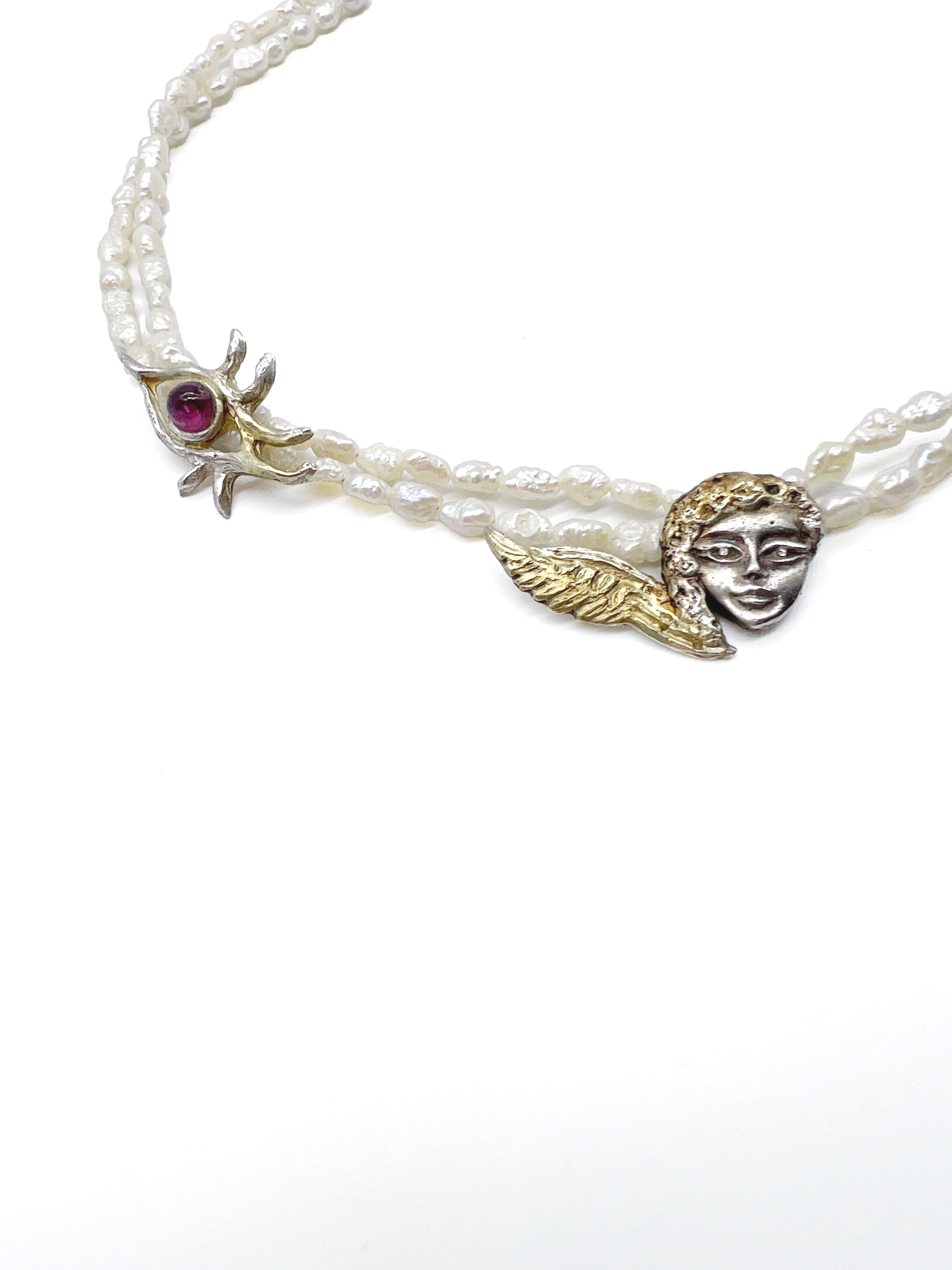 A pearl necklace with a decorative metal pendant featuring a female face and gold-colored wing detail, against a white background.