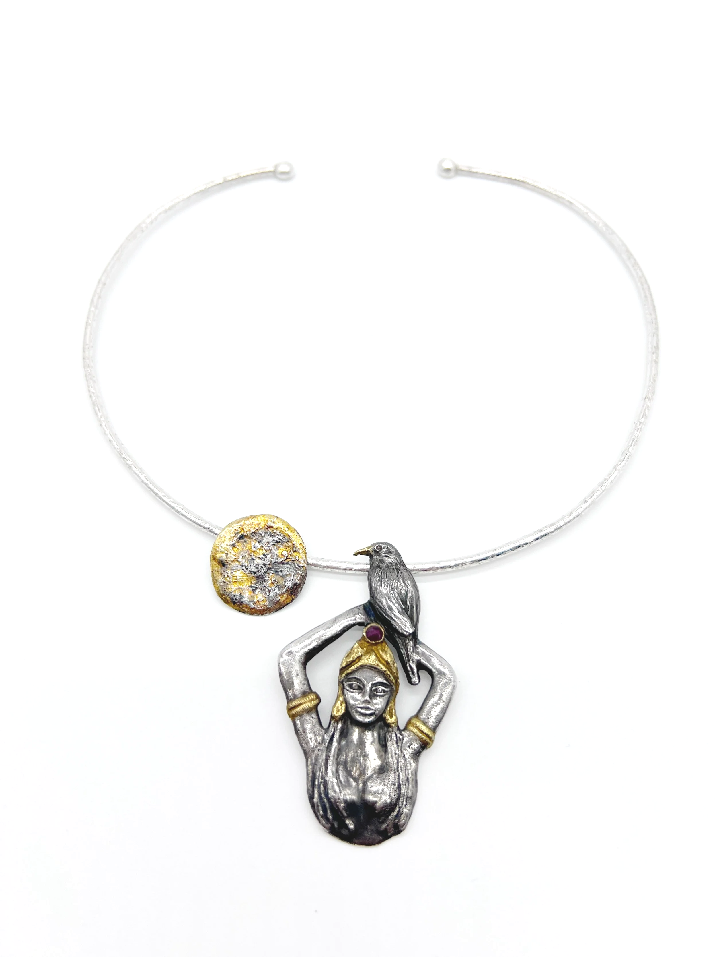 A silver necklace with a pendant of a woman with a gold headband and earrings, holding her hair up, with a bird and a textured gold circular element nearby.