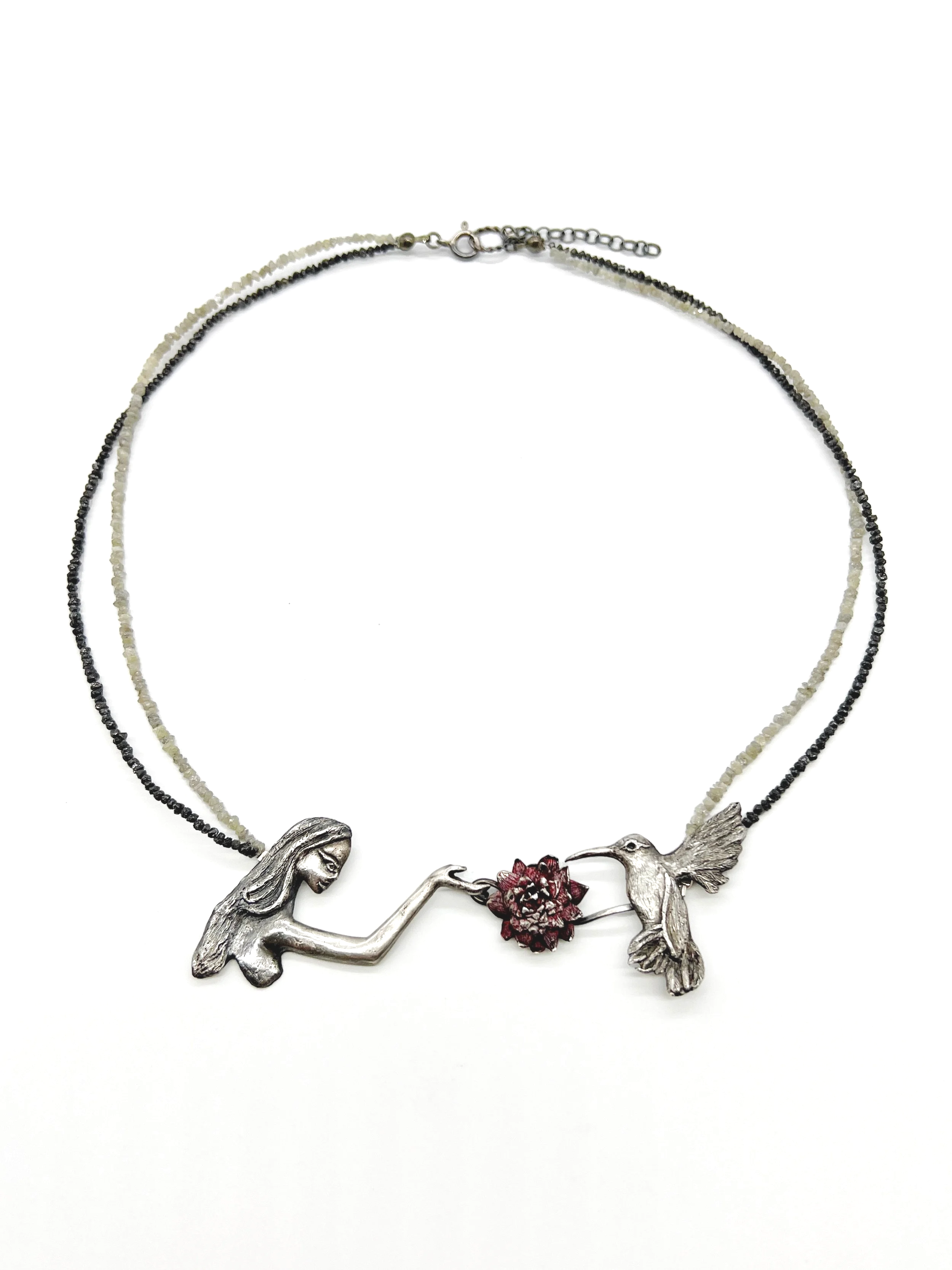 A necklace featuring a silver female figure with long hair, a hummingbird, and a red flower, all connected by a black and white beaded chain.