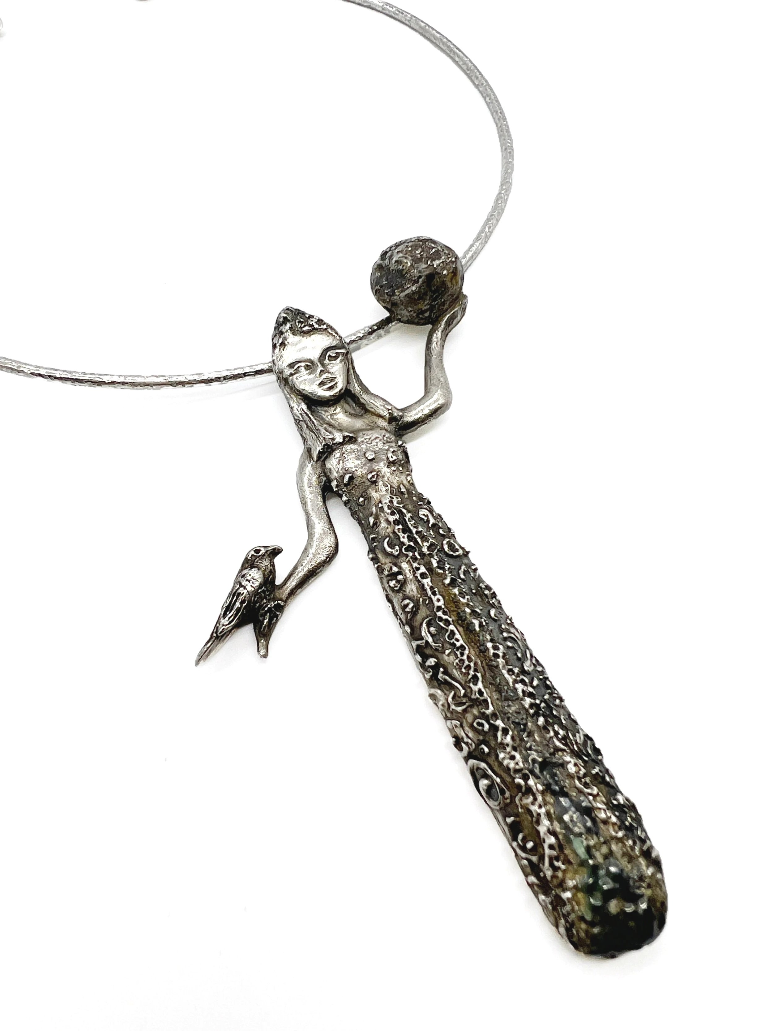 A silver jewelry necklace with a detailed mermaid figure holding a ball, decorative elements, and a bird hanging from the mermaid's tail against a white background.