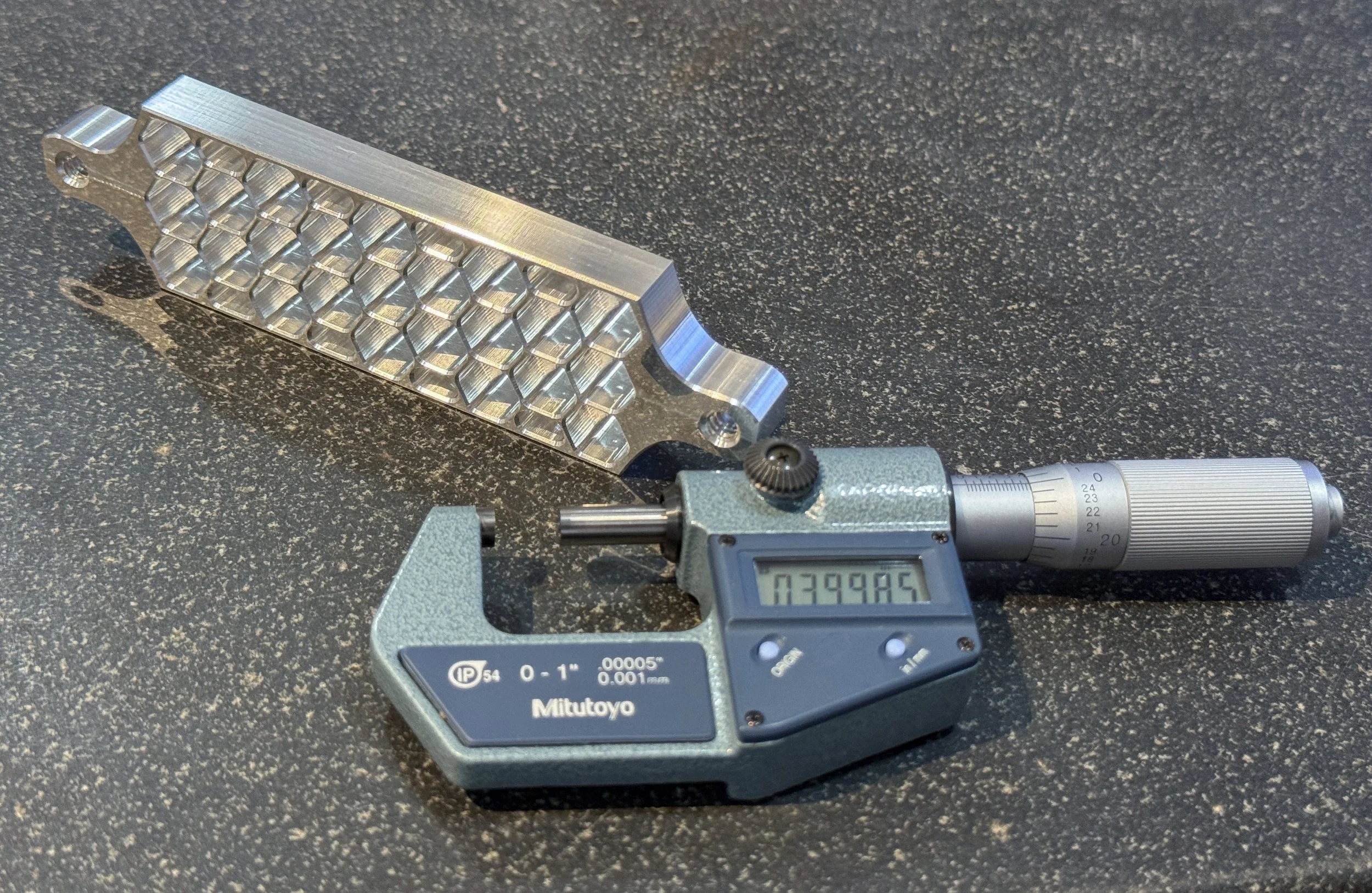 Machined aluminum honeycomb part beside a digital micrometer on a granite surface plate.