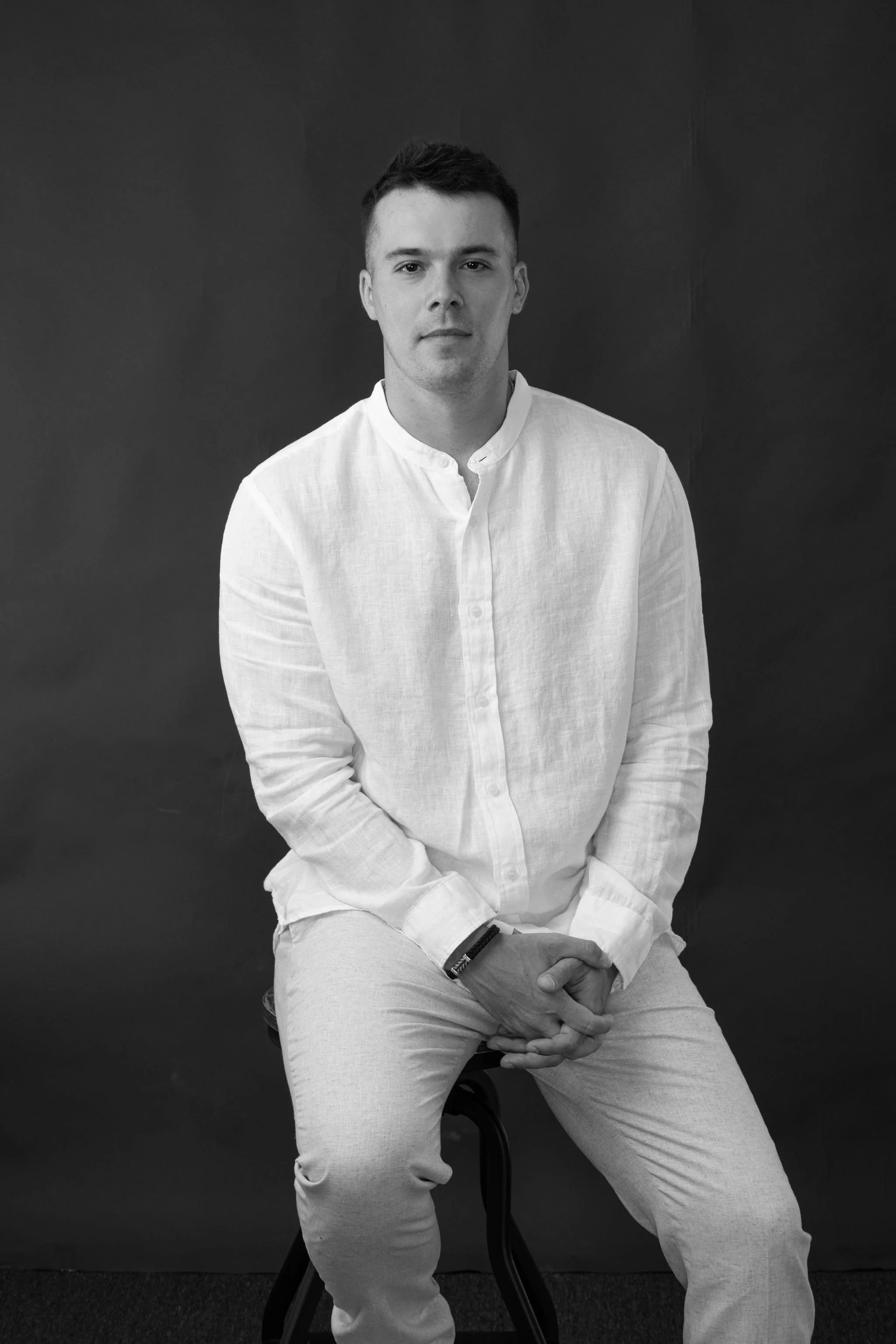 Black and white portrait of Grayson, wearing a white button-up shirt and light-colored pants, sitting on a stool against a dark background.