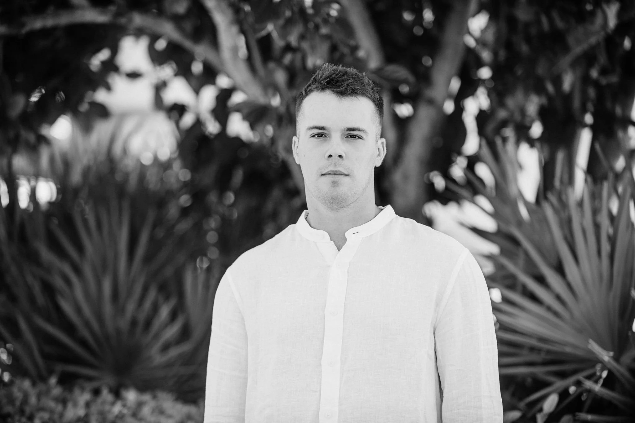 A headshot of Grayson, wearing a light-colored button-up shirt, standing outdoors in front of large plant leaves and trees.