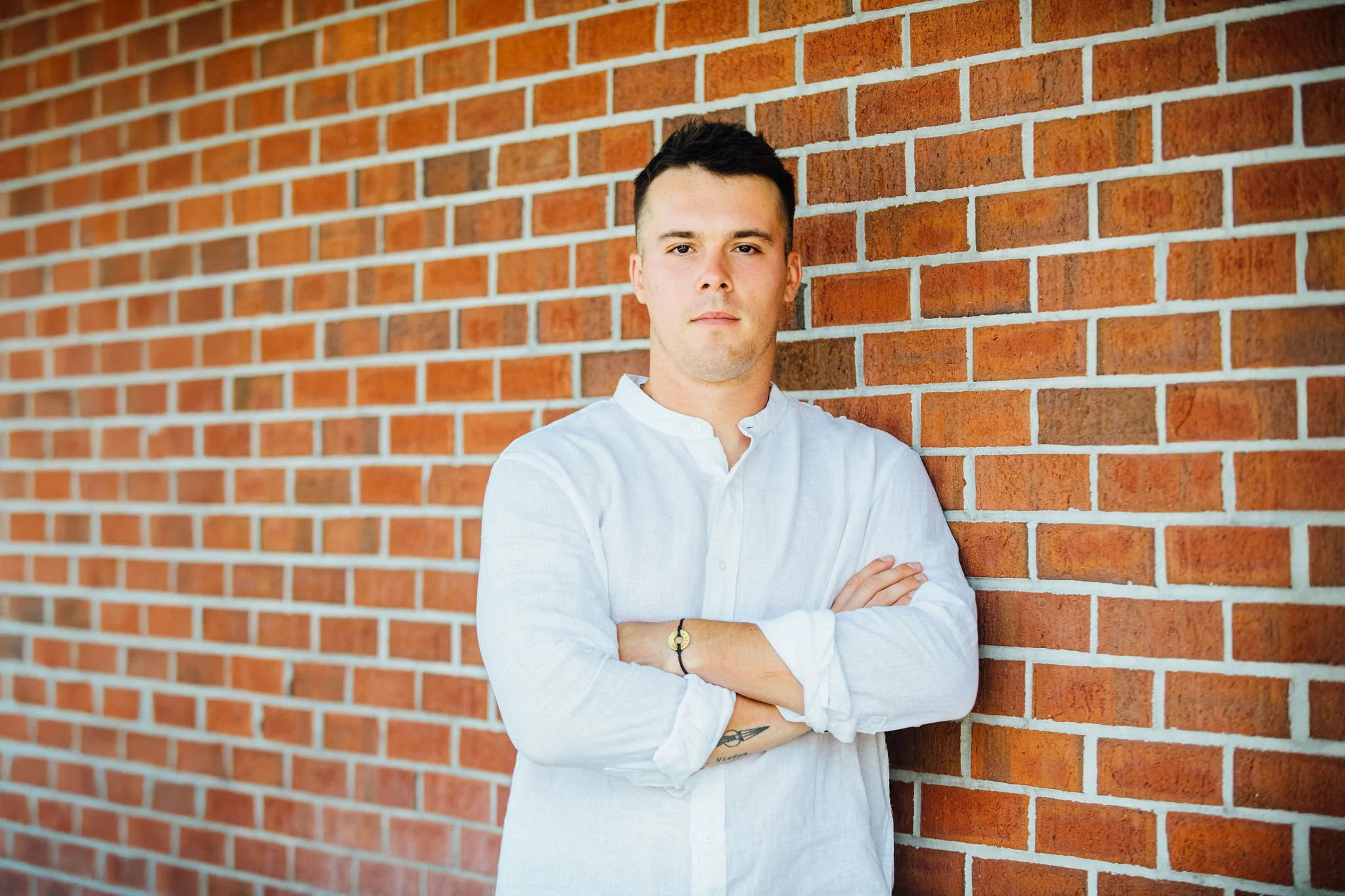 A headshot of Grayson McGraw, wearing a white shirt, standing with arms crossed against a red brick wall.