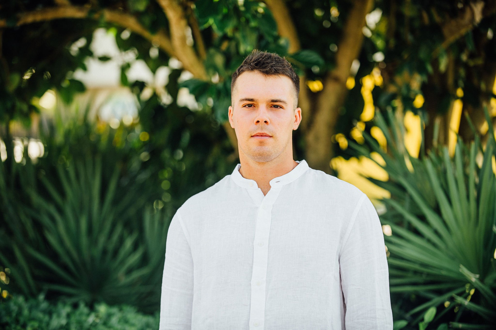 A headshot of Grayson wearing a white button-up shirt standing outdoors amidst green foliage and trees.