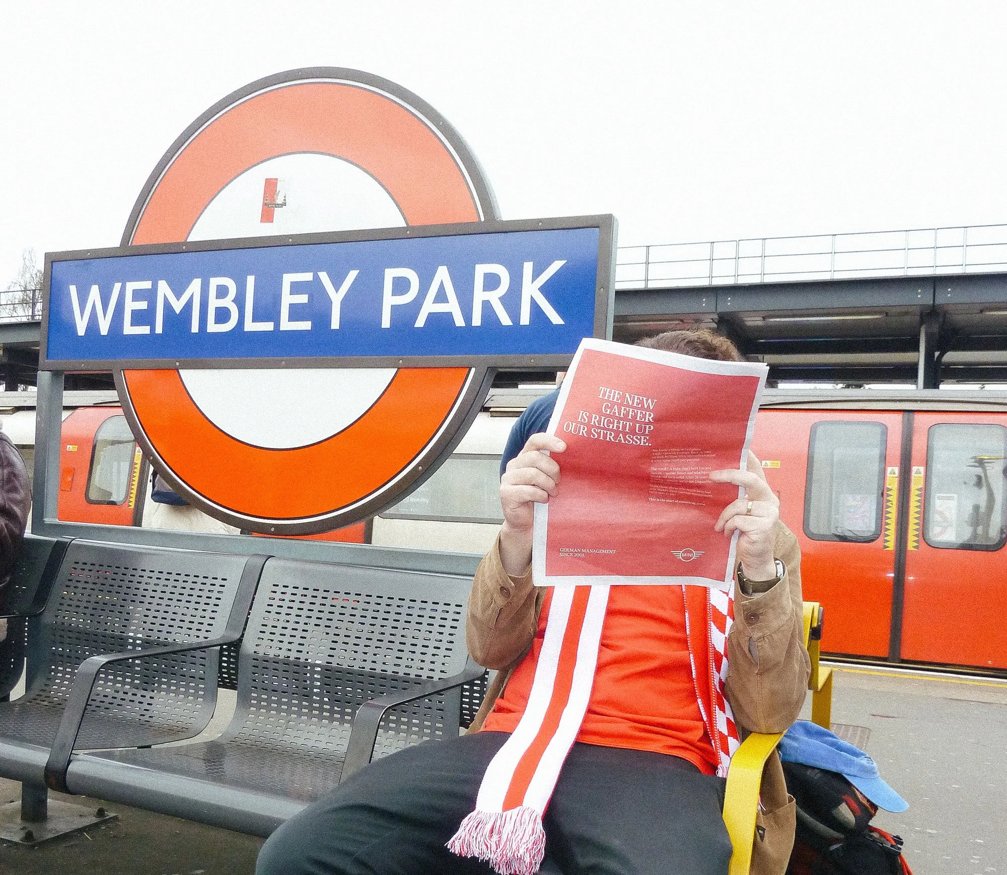 Person holding a red flyer at Wembley Park train station with a Timpson sign and orange train in the background.