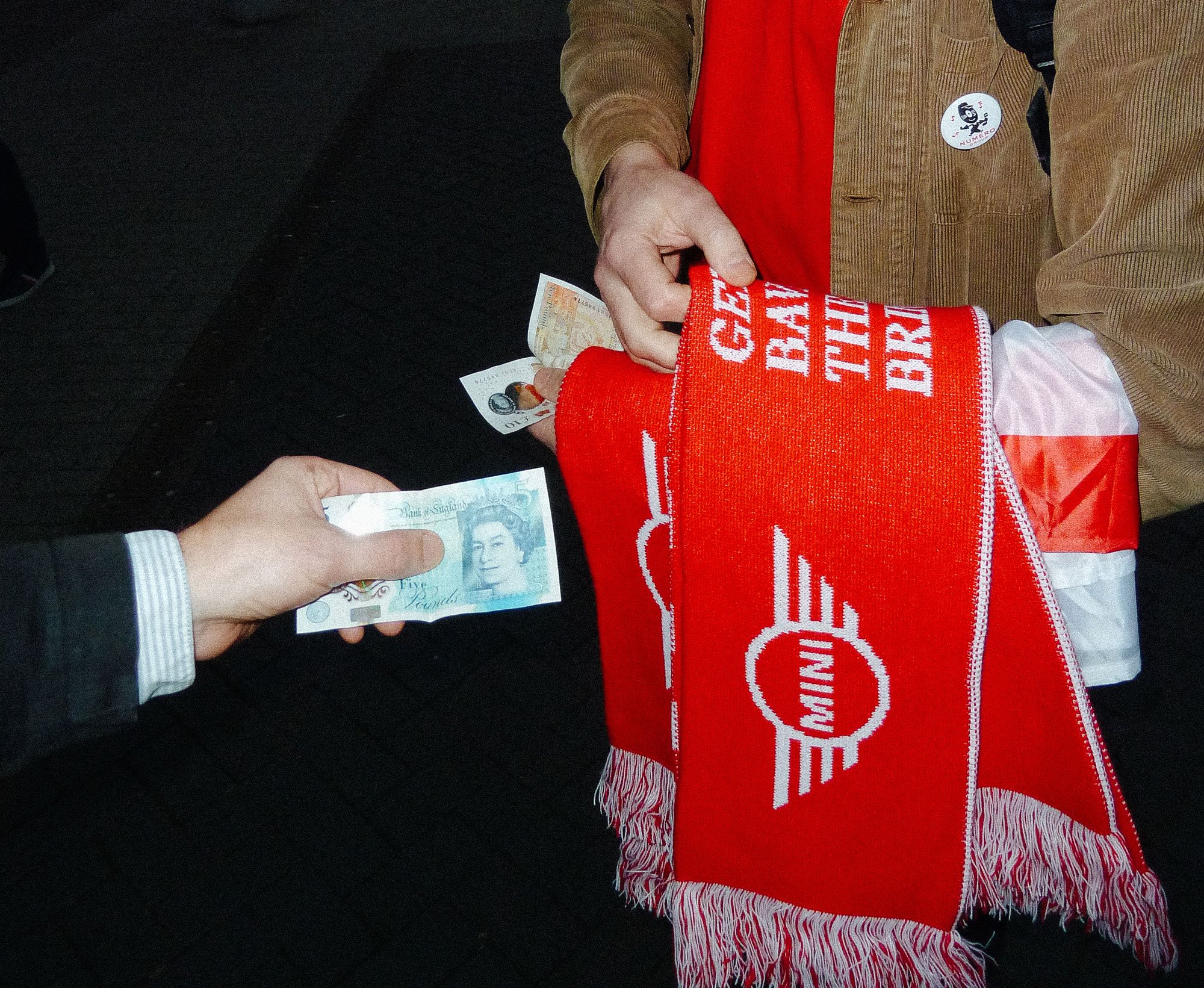 A person dressed in a red shirt and tan jacket is holding a 5-dollar bill, with a red and white scarf displaying the words 'Get Back to Work'. There is a round button with a smiling cartoon character on the jacket.