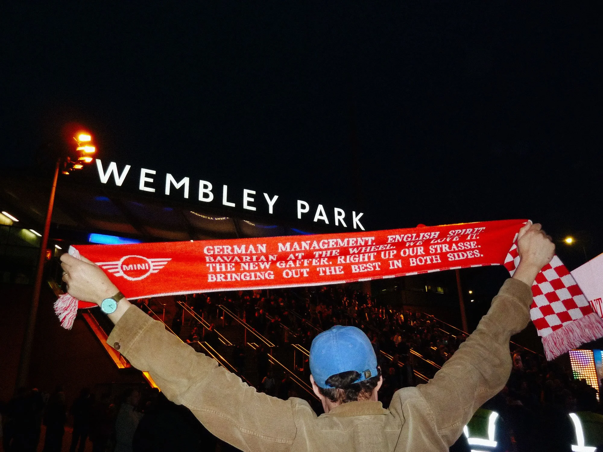 People in a stadium holding a red scarf that reads, 'GERMAN MANAGEMENT FILM AT THE WHEEL! BRING OUT THE BEST IN BOTH SIDES.' The stadium is called Wembley Park, with a large sign visible in the background; the scene is at night.