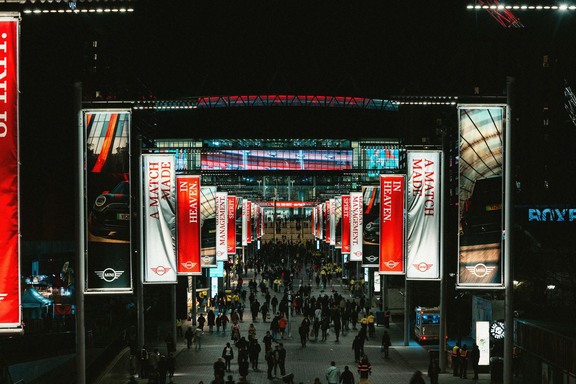 Night view of a bustling city street leading to a large illuminated building with banners that read 'A Match in Maden.' People are walking along the street, and cars are parked nearby. The building has large glass windows and several digital screens.