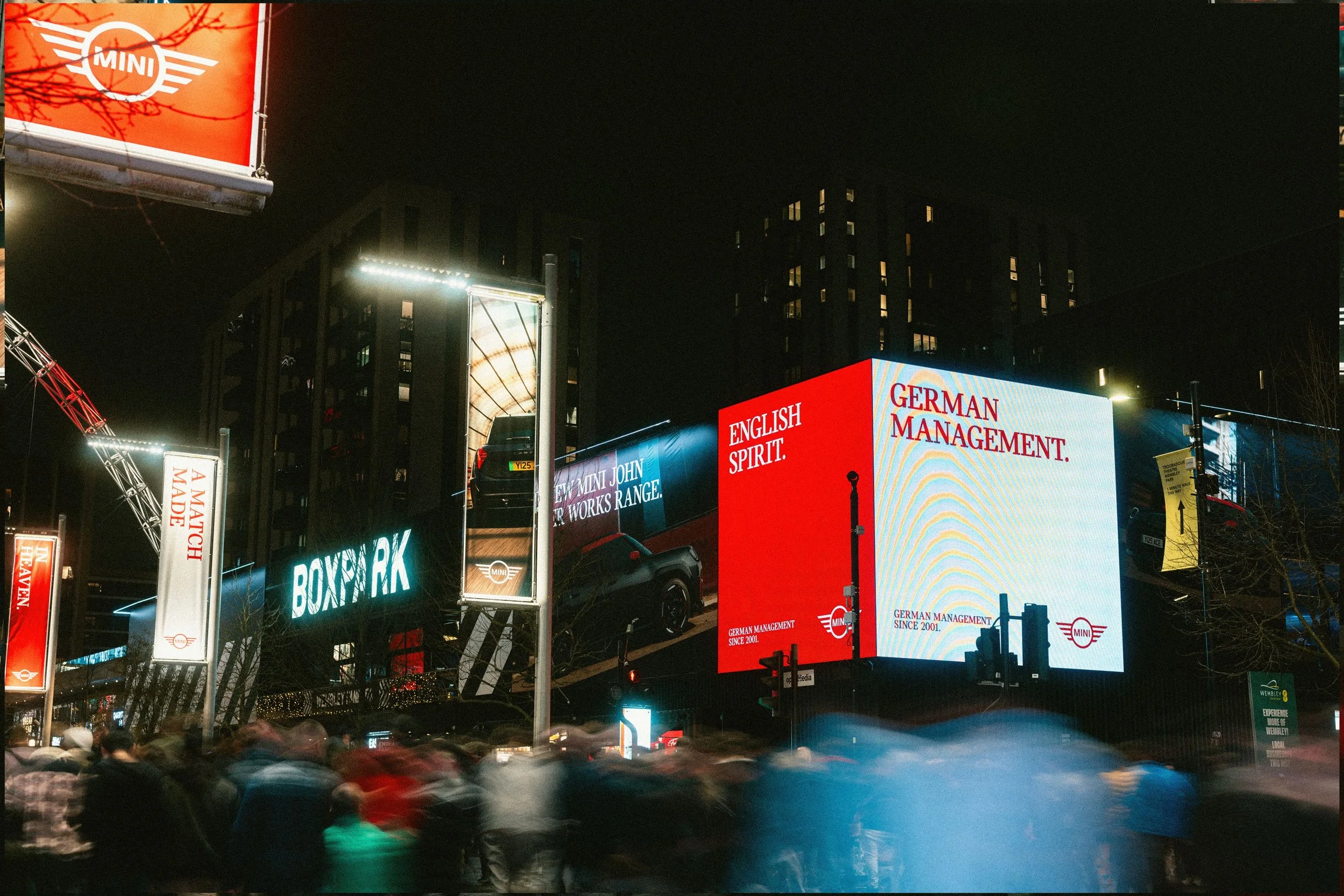 Night scene of Times Square with illuminated billboards and signs, including advertisements for Mini cars, German management, and Broadway, with a crowd of people and blurred motion of vehicles.